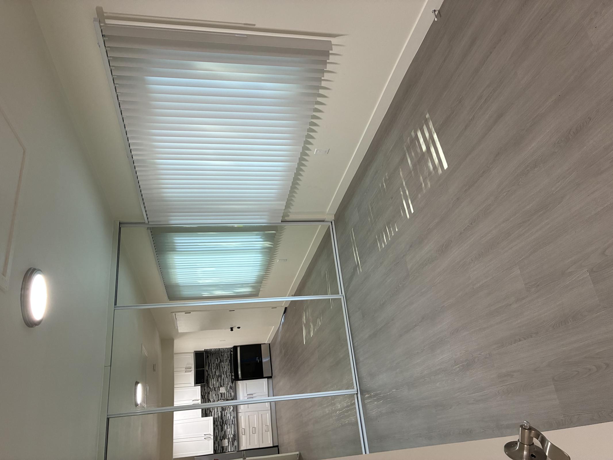 A bright, minimalist room featuring grey laminate flooring. On one side, there’s a large mirrored closet with sliding doors. A window with vertical blinds allows natural light to enter, and in the background, a modern kitchen is visible with white cabinetry and a dark stove. The atmosphere is clean and inviting.