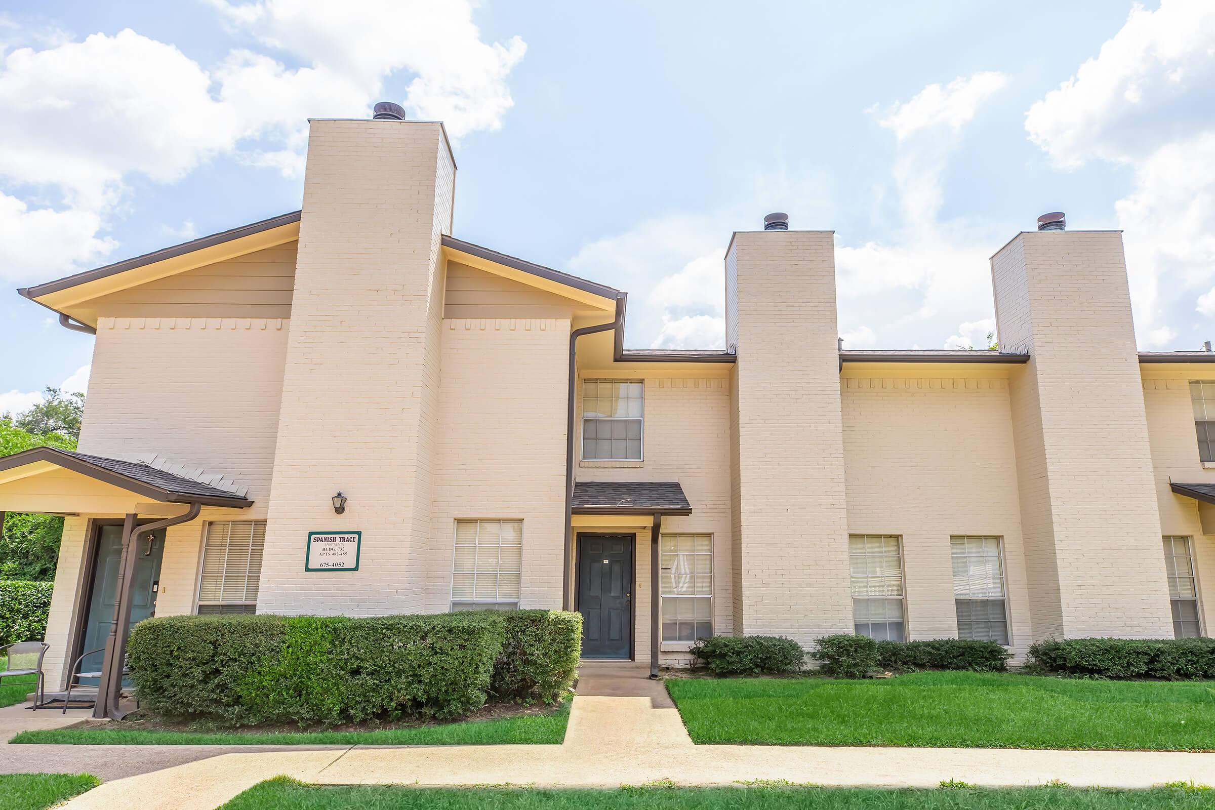 Two-story residential building with a light-colored brick exterior. The front features a gray door and multiple windows. There are two tall chimneys on the roof and neatly trimmed hedges lining the walkway. The sky is partly cloudy, giving a bright and inviting atmosphere.