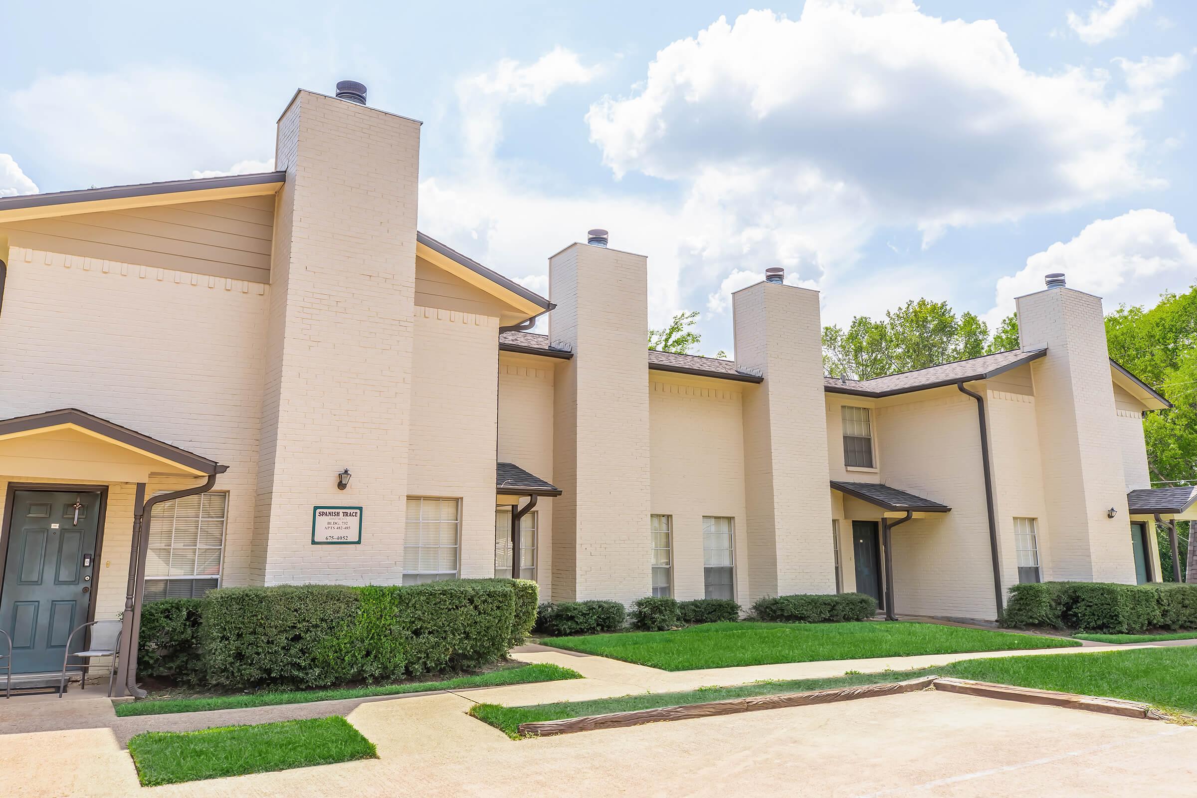 A two-story cream-colored building with multiple chimneys, surrounded by manicured green lawns and shrubs. The structure features large windows and a dark roof, with a paved area in front. The sky is partly cloudy, adding a bright ambiance to the scene.