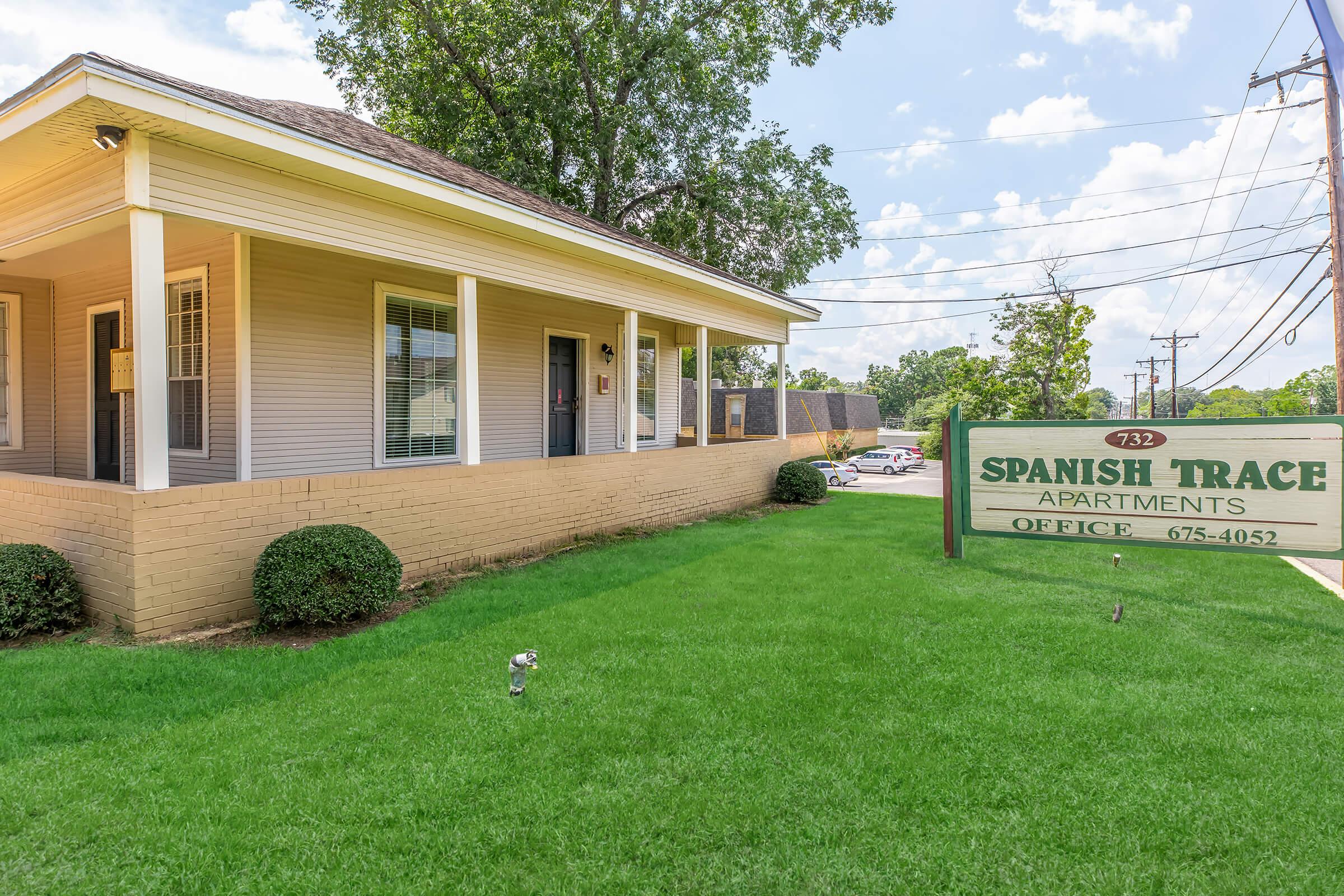 A view of the Spanish Trace Apartments office building, featuring a light-colored, one-story structure with a porch, surrounded by green grass and landscaped bushes. A sign indicating "Spanish Trace Apartments" and the office number is prominently displayed in front, under a blue sky with scattered clouds.