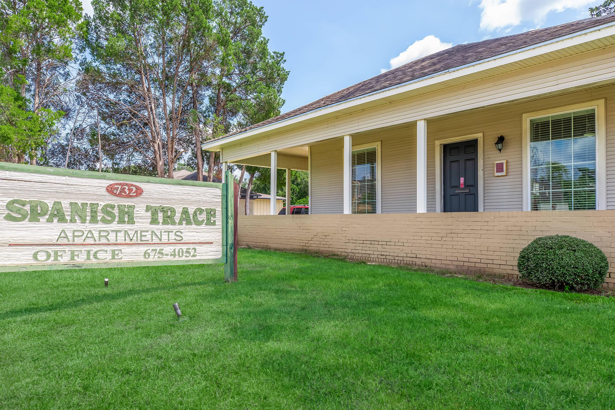 Sign for Spanish Trace Apartments in front of a light-colored building with a porch. The office number is displayed as 732, and the contact number is 675-4052. The surrounding area features green grass and trees, with a clear blue sky above.
