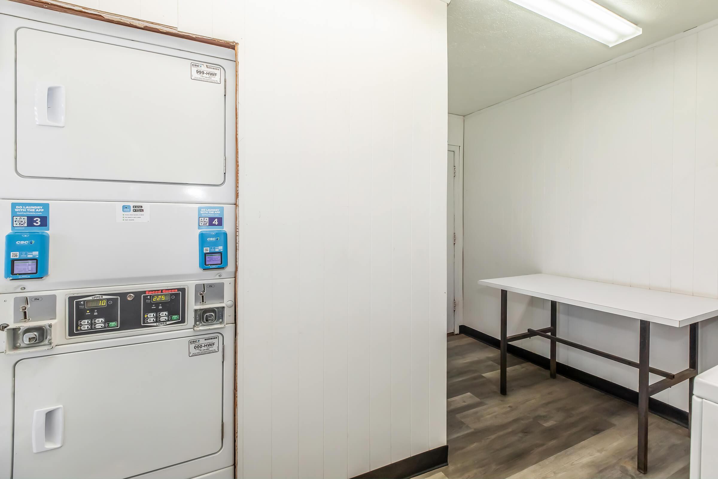 A clean and modern laundry room featuring stacked washer and dryer units on the left, with control panels visible. To the right, there is a simple white table against a plain wall. The flooring is a neutral tone, adding to the tidy and organized appearance of the space.