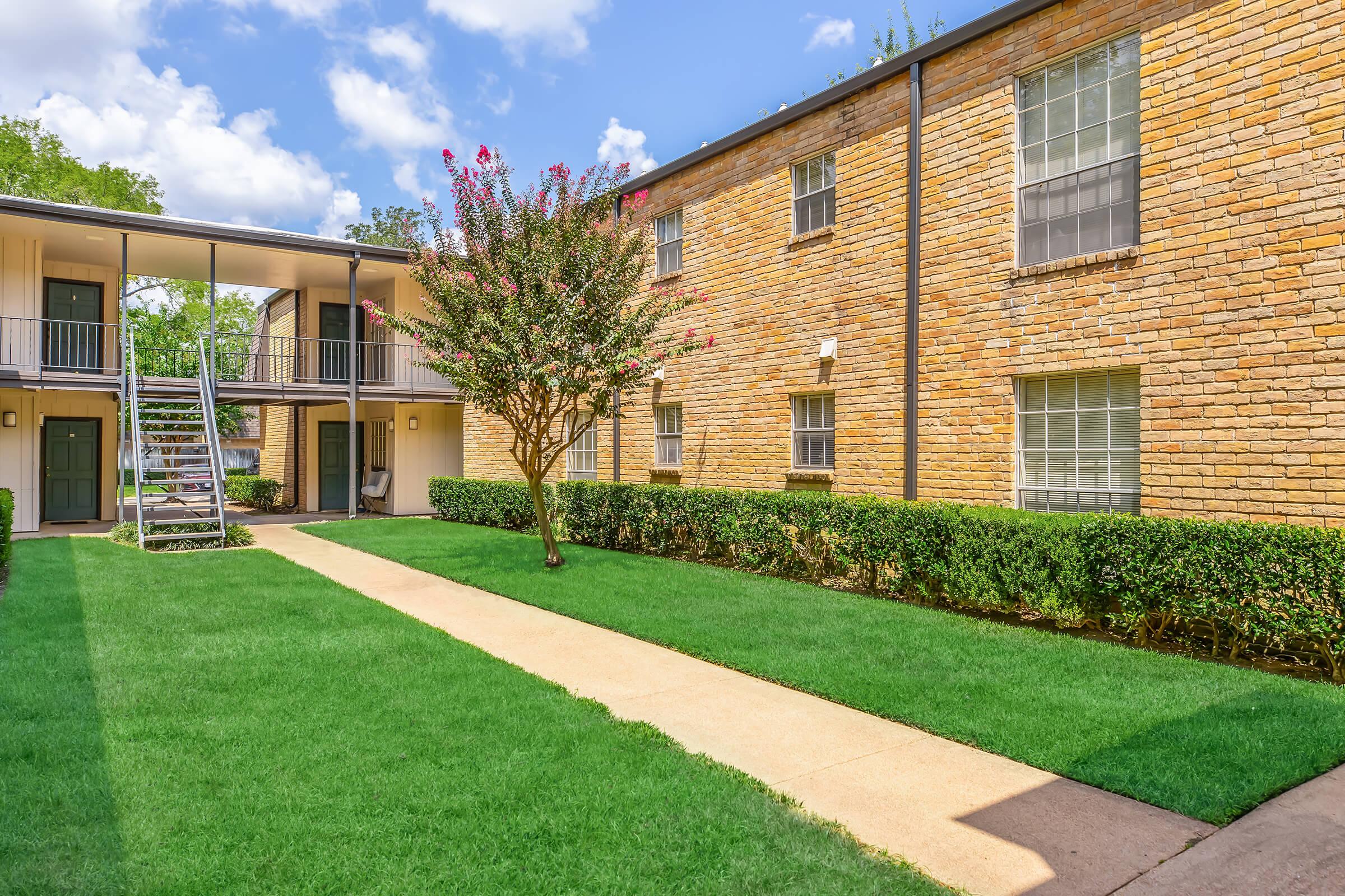 A well-maintained apartment complex featuring two buildings made of brick, surrounded by lush green grass. A paved walkway leads through the courtyard, with a flowering tree and a staircase visible. The sky is bright with scattered clouds, adding to the inviting atmosphere.