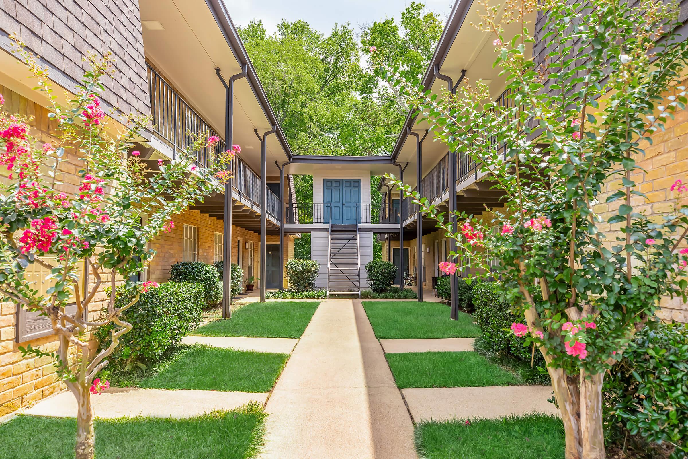 A well-maintained courtyard of an apartment complex featuring lush green grass, flowering bushes, and a central staircase leading to upper-level units. The layout includes two rows of buildings with balconies, surrounded by vibrant greenery and colorful blossoms, creating a welcoming atmosphere.