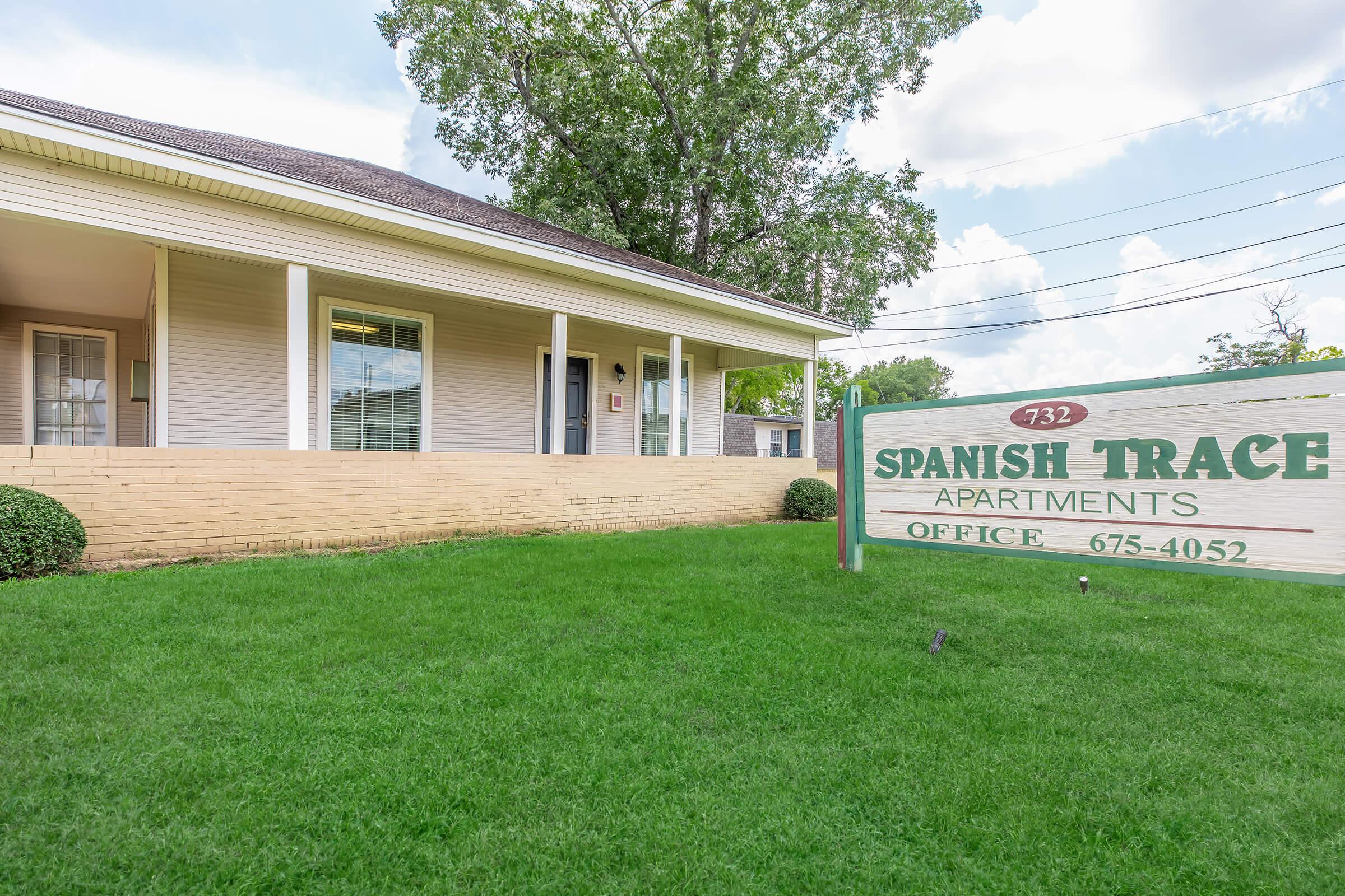 A view of the Spanish Trace Apartments, featuring a light-colored building with a front porch and a sign displaying the name and office number. The landscape includes a well-maintained green lawn and nearby shrubs, with a clear sky in the background.