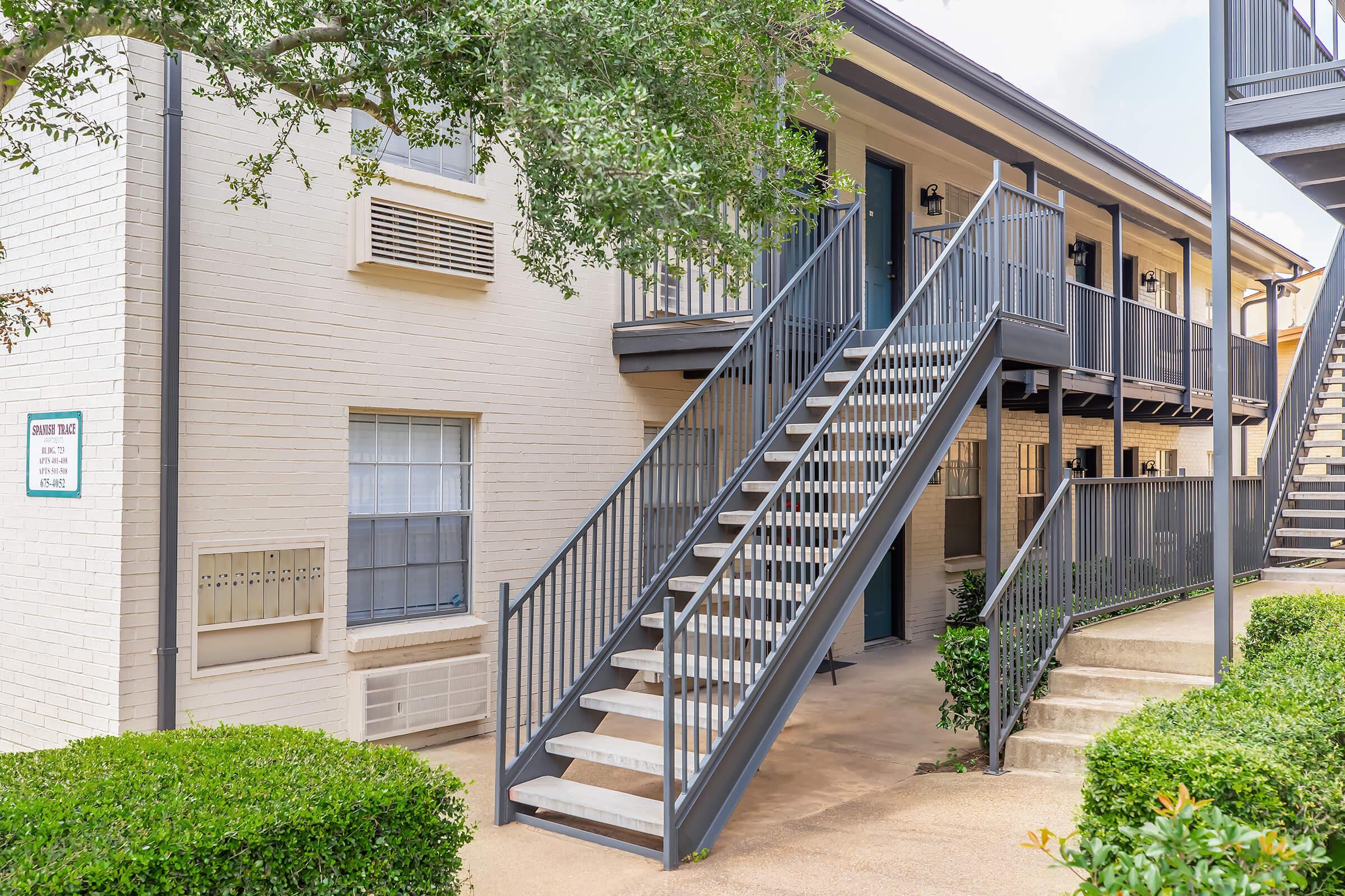 Exterior view of an apartment building featuring two levels. A set of stairs leads to the upper floor, with a pathway and landscaped shrubs in the foreground. Sunlight casts shadows on the building's beige walls, and green foliage adds to the inviting atmosphere.