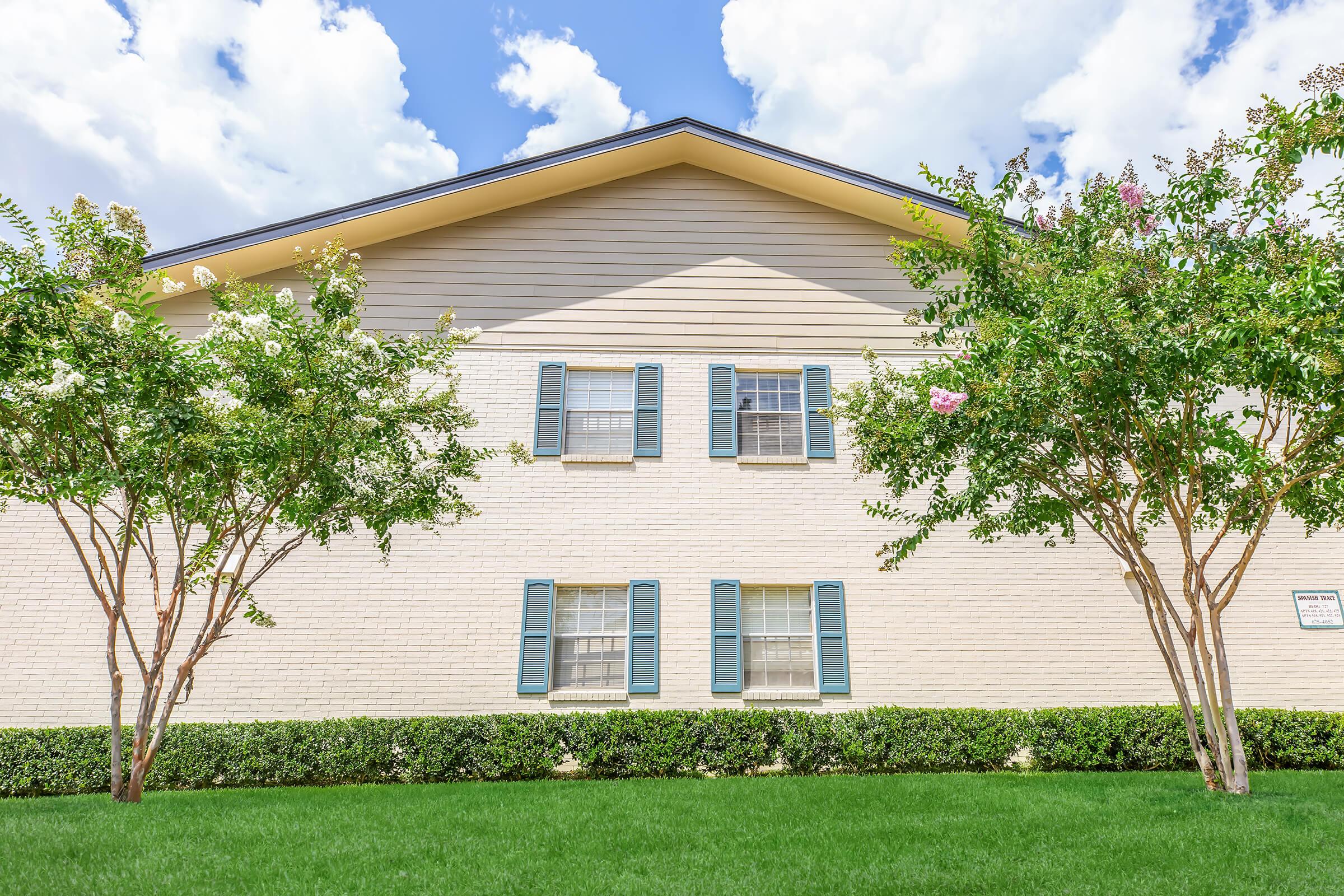 A brick building with light-colored siding and blue shutters. It features three windows on the upper level and is surrounded by well-manicured green grass, small bushes, and flowering trees under a blue sky with fluffy white clouds.