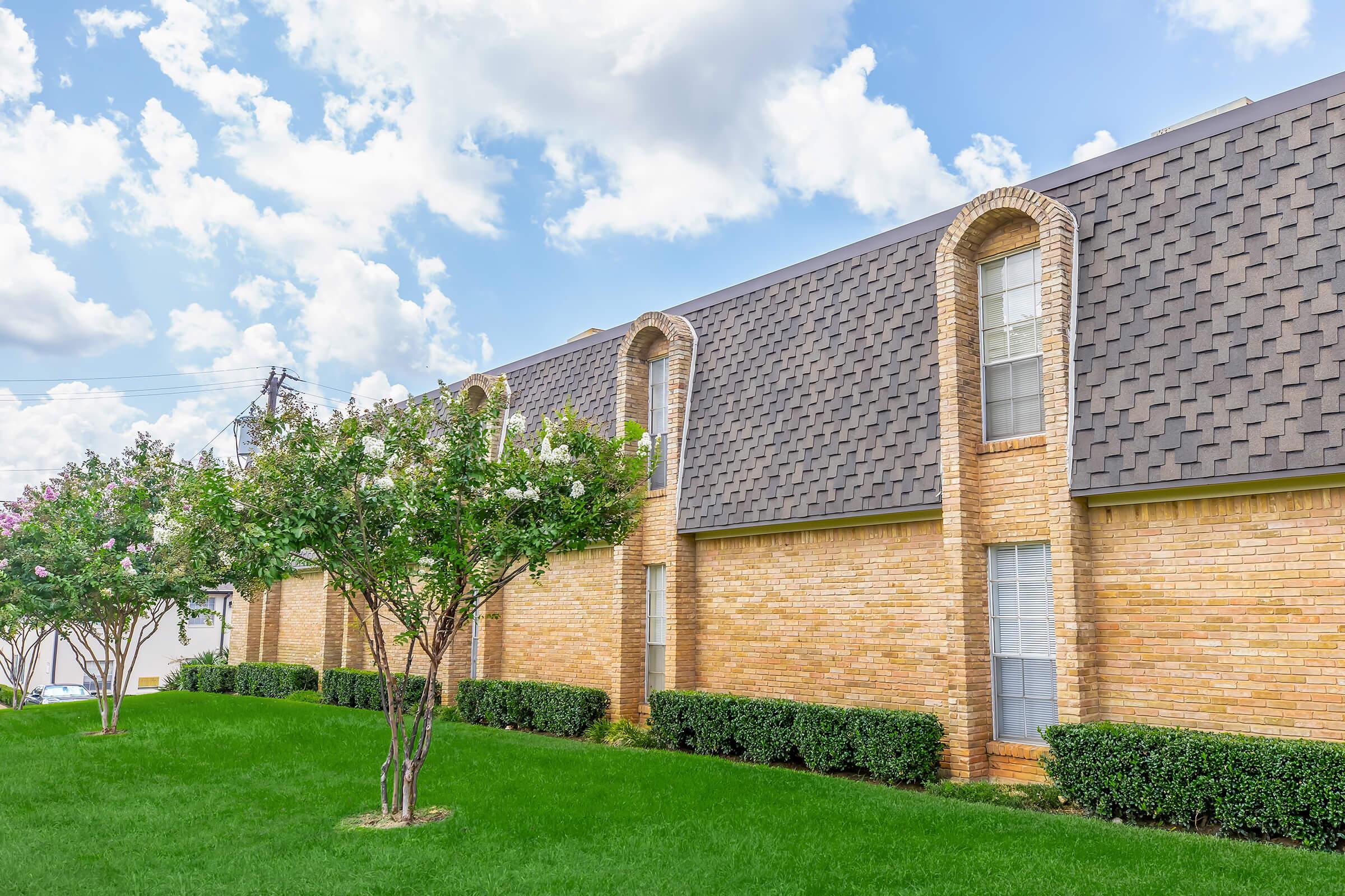 A modern brick building with a textured roof, featuring large windows and decorative archways. The structure is surrounded by lush green grass and flowering trees under a partly cloudy blue sky.
