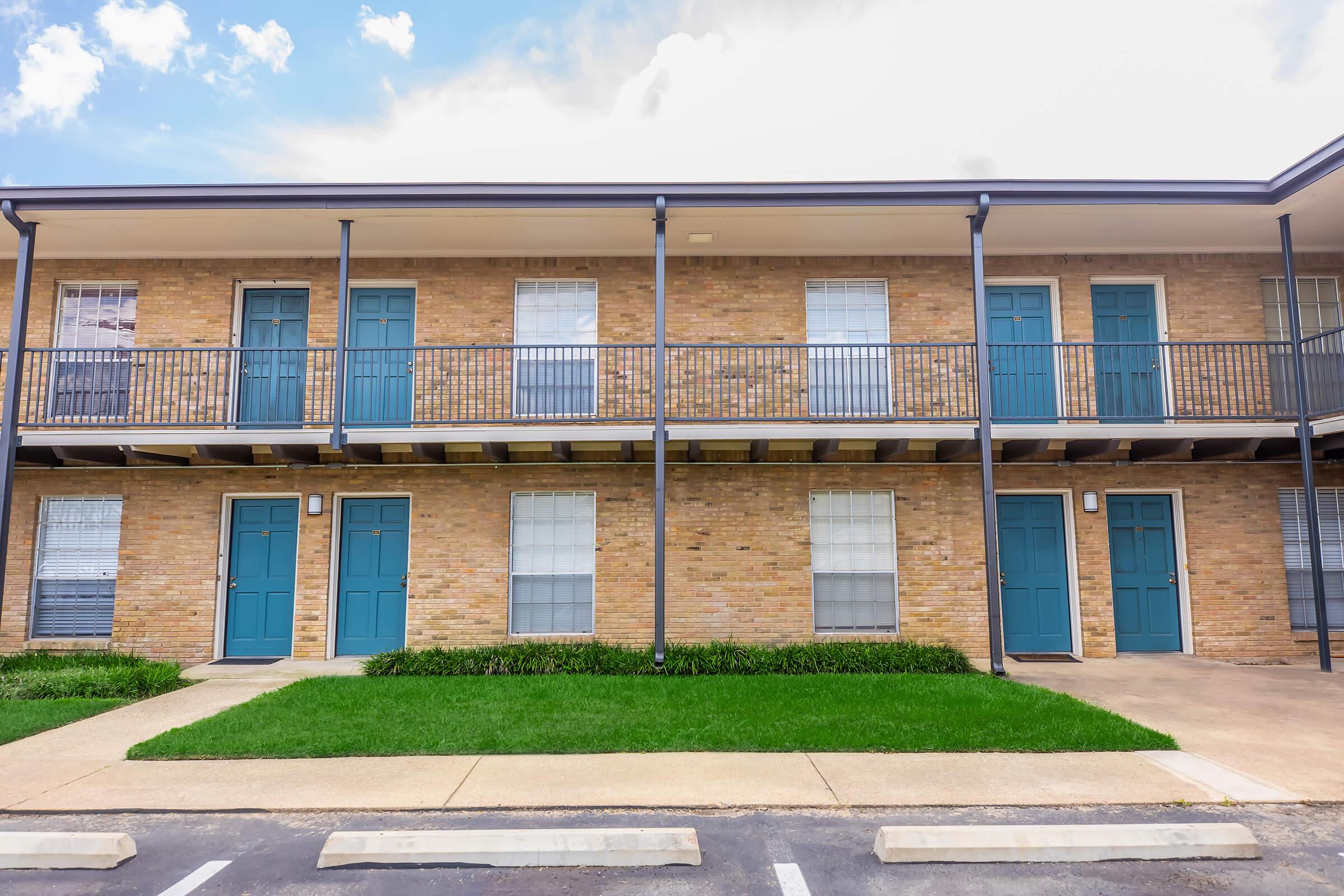 Two-story brick apartment building with turquoise doors. Each level features a balcony with metal railings, and there are several windows on both floors, all framed with white blinds. A neat green lawn runs along the front of the building, and a paved parking area is visible in the foreground.