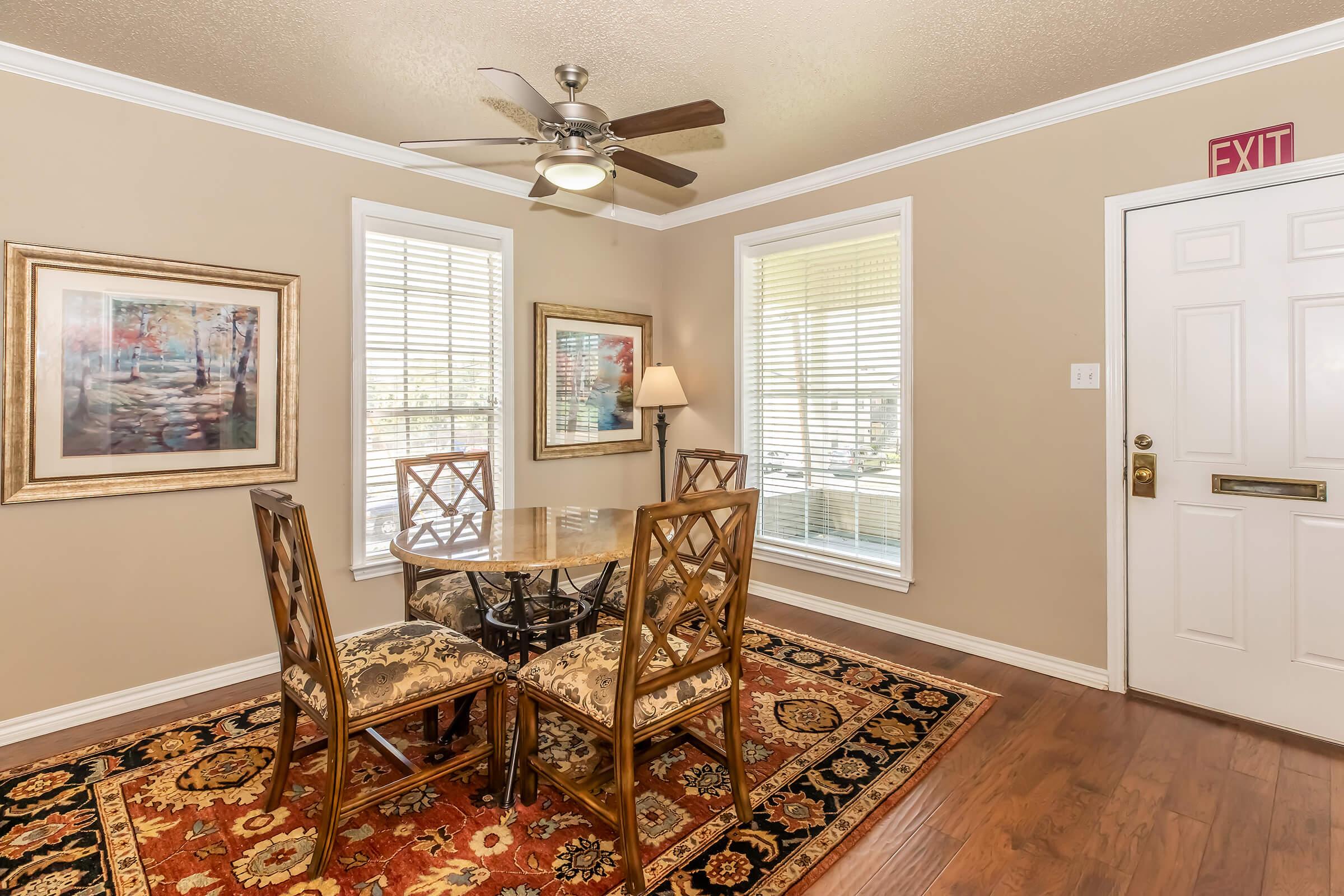 A well-lit dining area featuring a round table with four decorative chairs, set on a vibrant area rug. Two windows allow natural light to fill the space, while framed artwork adorns the walls. A ceiling fan provides airflow, and an exit sign is visible near the door.