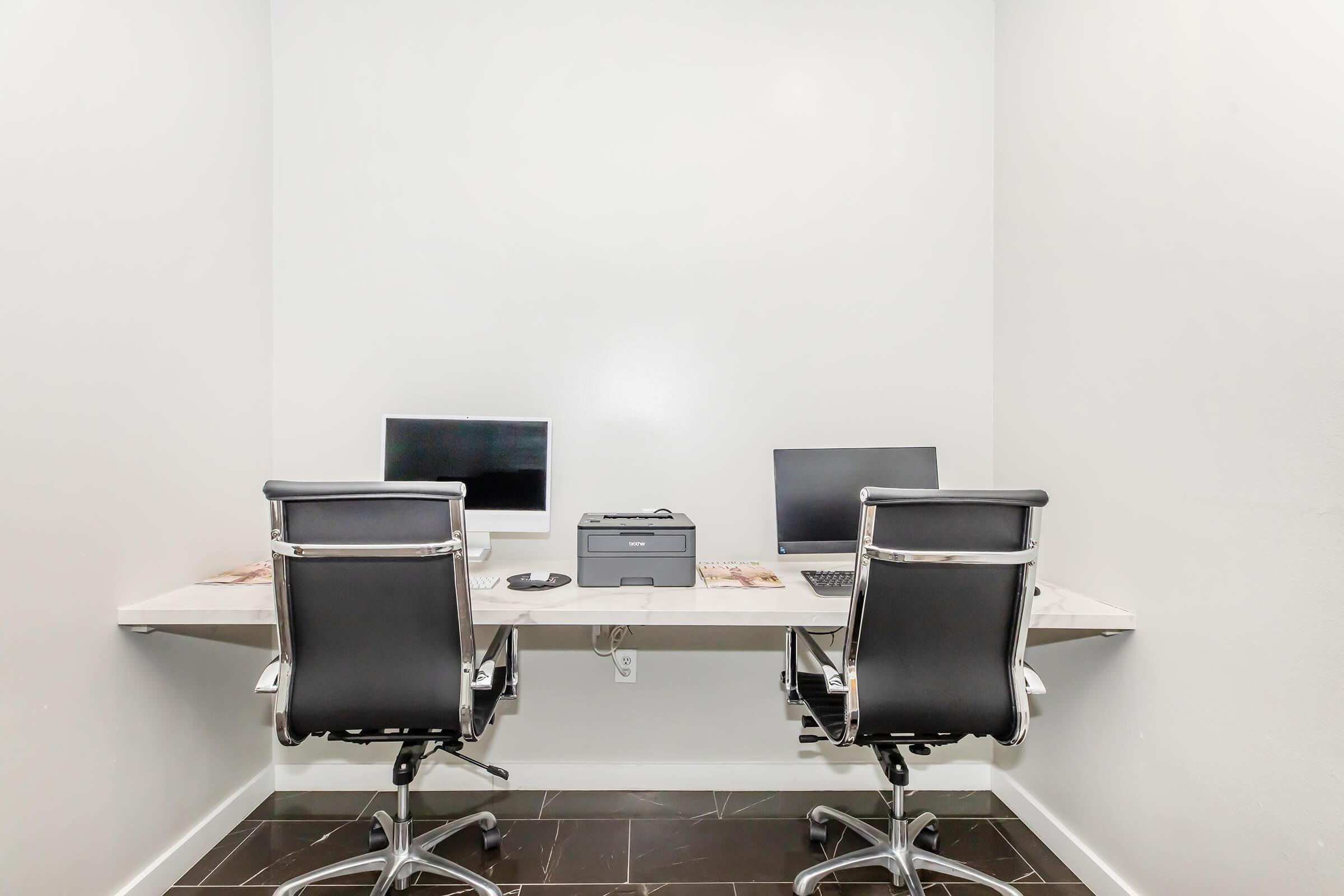 A minimalist office workspace featuring two sleek, black swivel chairs at a white wall-mounted desk. Each chair is positioned in front of a computer monitor. A printer is placed on the desk, and the floor is covered with dark tiles. The walls and decor are simple and modern, creating a clean, organized environment.