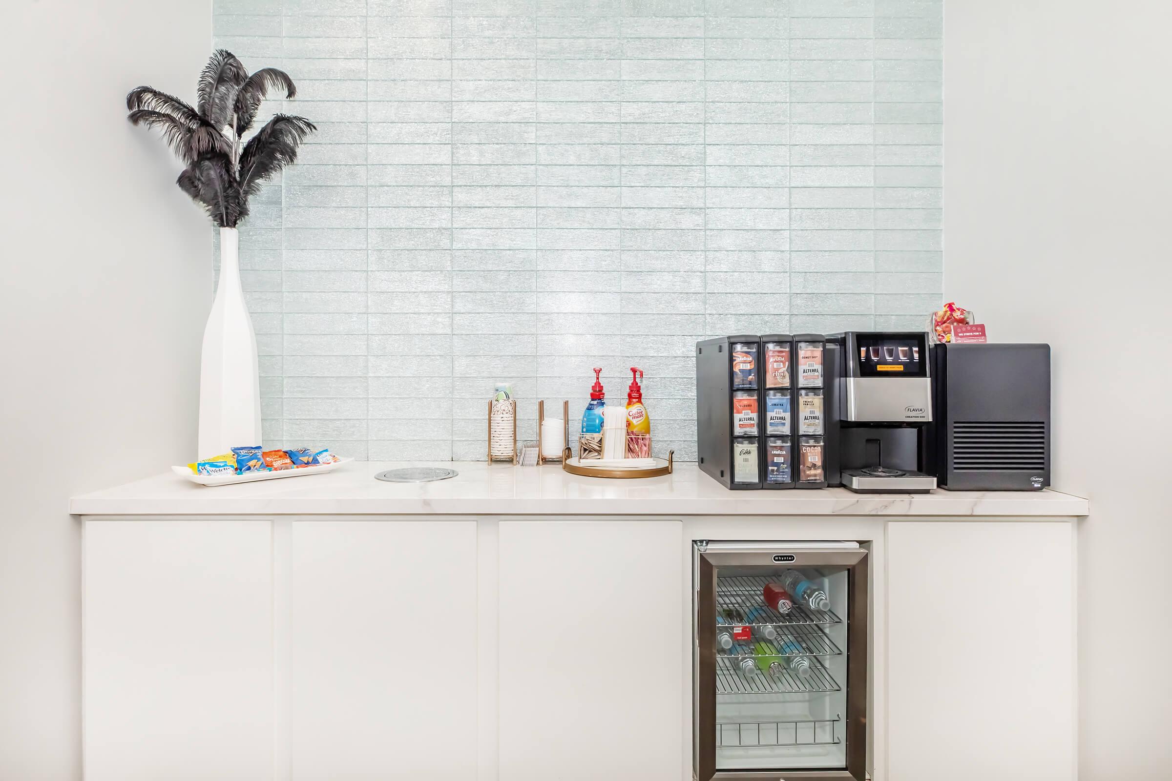 Modern kitchenette featuring a white cabinet with a small fridge displaying drinks, a coffee machine with various flavor syrups, and a decorative vase with feathers. Snacks are arranged on the countertop beside a dispenser. The backsplash is light blue tiled for an elegant touch.