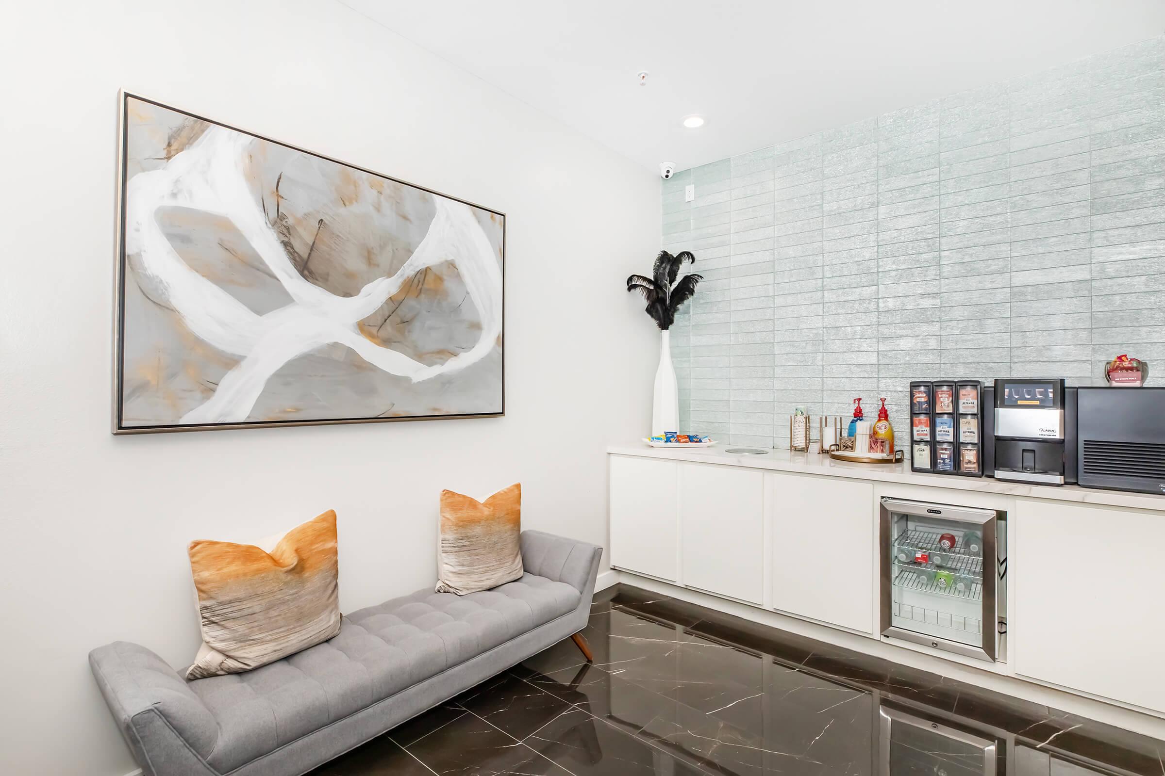 A modern lounge area featuring a gray sofa with decorative pillows, a sleek white cabinet, a small refrigerator, and a coffee station. The wall displays an abstract artwork with neutral tones. The floor has dark tiles, and there is a decorative plant in a white vase, creating an inviting atmosphere.