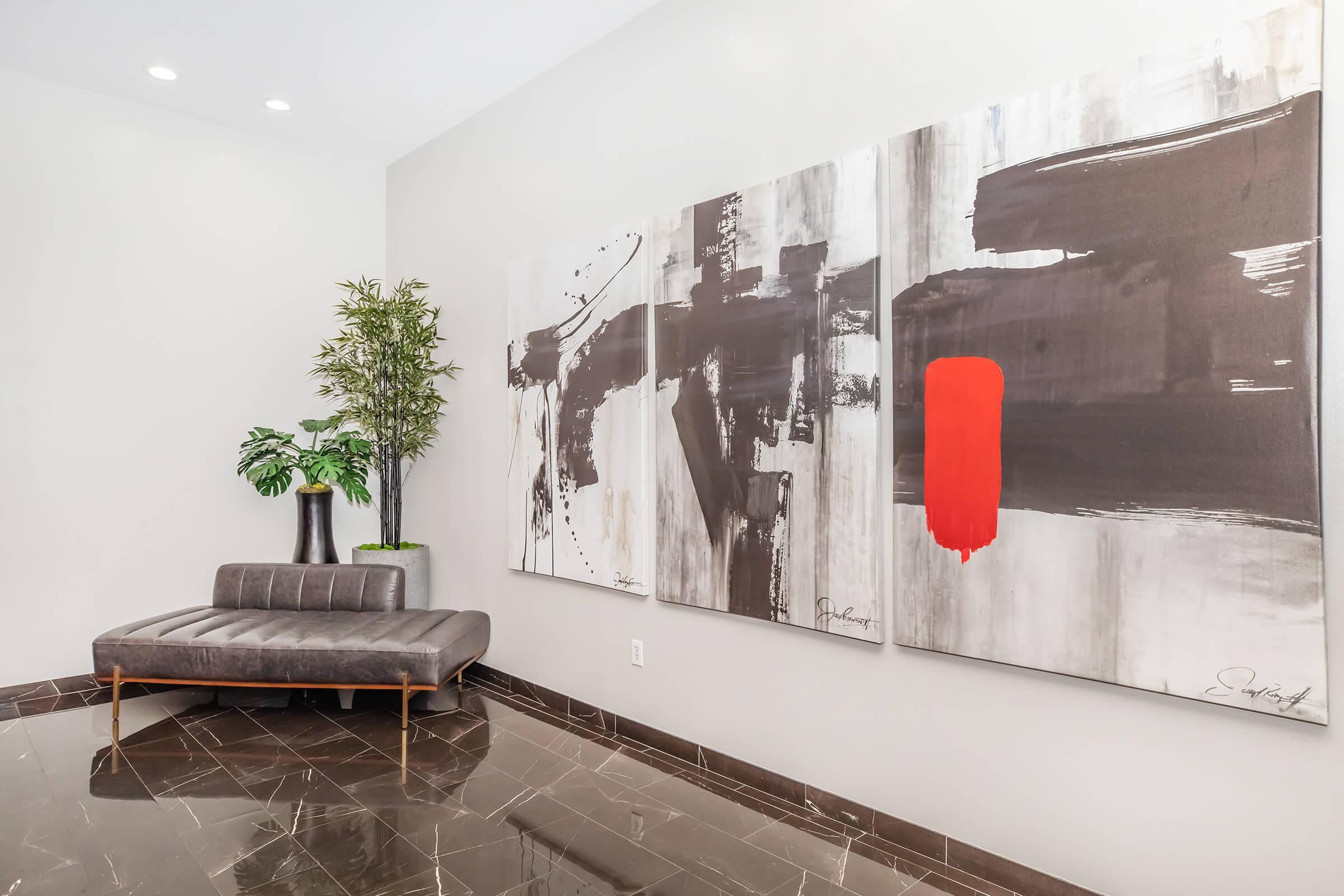 A modern interior featuring a sleek gray couch against a minimalist wall. Three large abstract paintings dominate the space, with bold black and white strokes and a striking red element. A tall green plant in a black vase adds a touch of nature to the contemporary setting, complemented by glossy dark flooring.