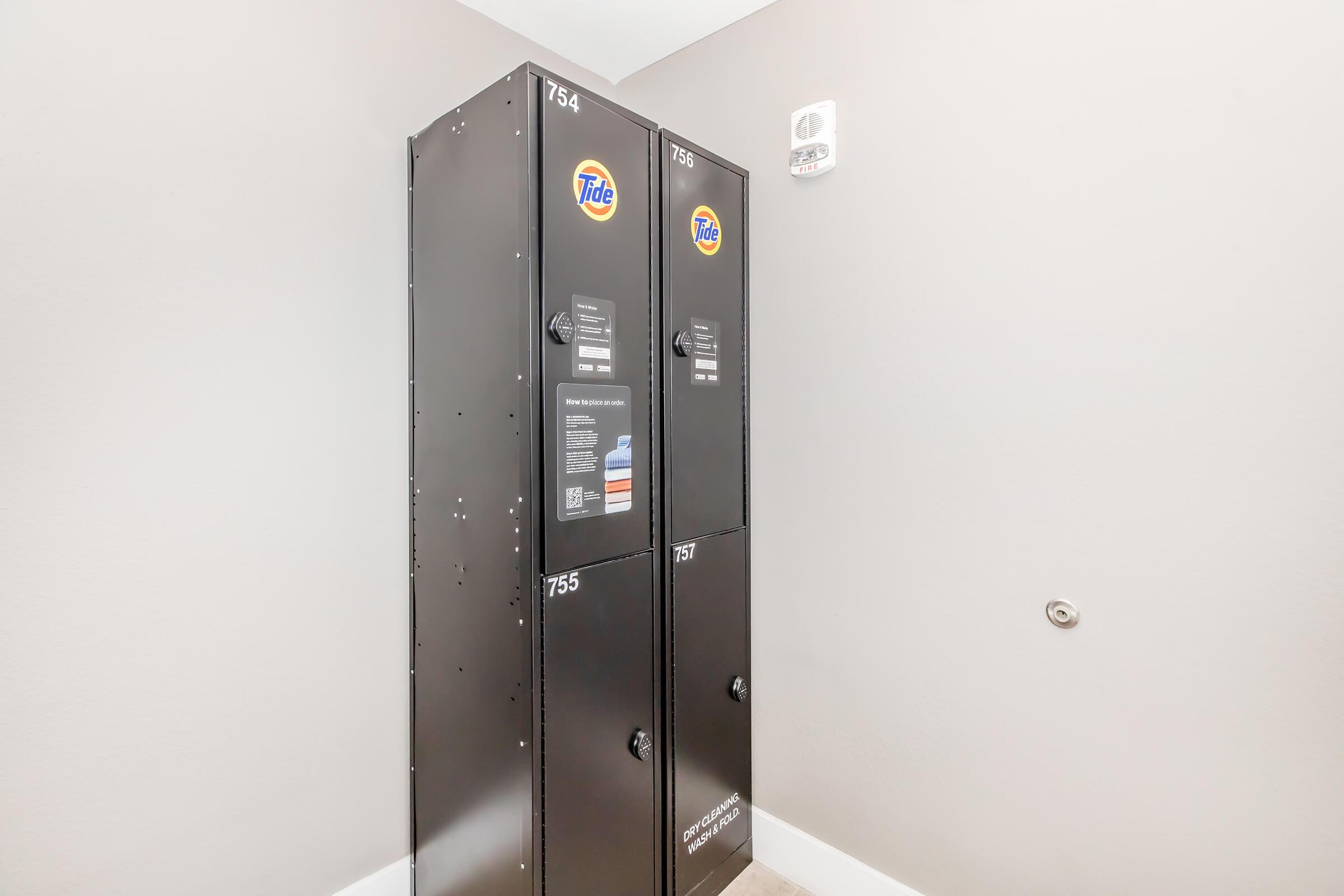 Two black laundry machines with Tide branding, positioned against a light gray wall. Each machine has a number, 754 and 755, displayed prominently. The area appears clean and well-maintained, with no visible clutter. There is a small white device or sensor mounted above the machines on the wall.