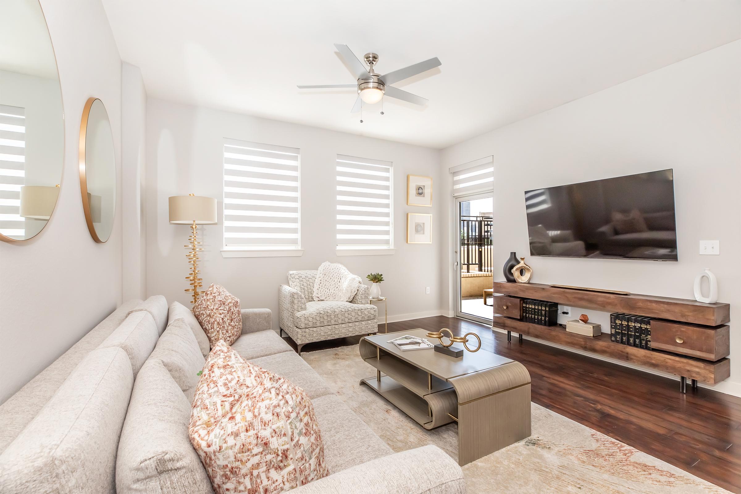 A modern living room featuring a light gray sectional sofa with decorative pillows, a round mirror on the wall, and a wooden TV stand with decorative items. A ceiling fan hangs above, and natural light streams in from large windows with dual-tone shades. A cozy armchair is positioned nearby, creating an inviting atmosphere.