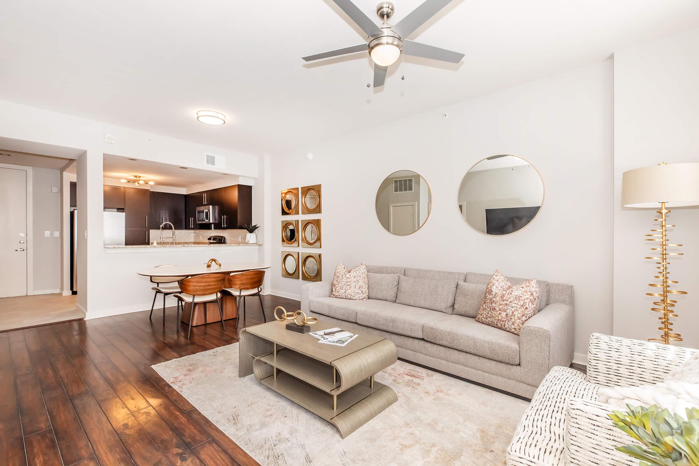 A modern living room featuring a light gray sofa with patterned pillows, a round coffee table, and a cozy area rug. Two circular mirrors adorn the wall, and a dining area with a wooden table and chairs is visible in the background. A stylish floor lamp adds warmth to the space.