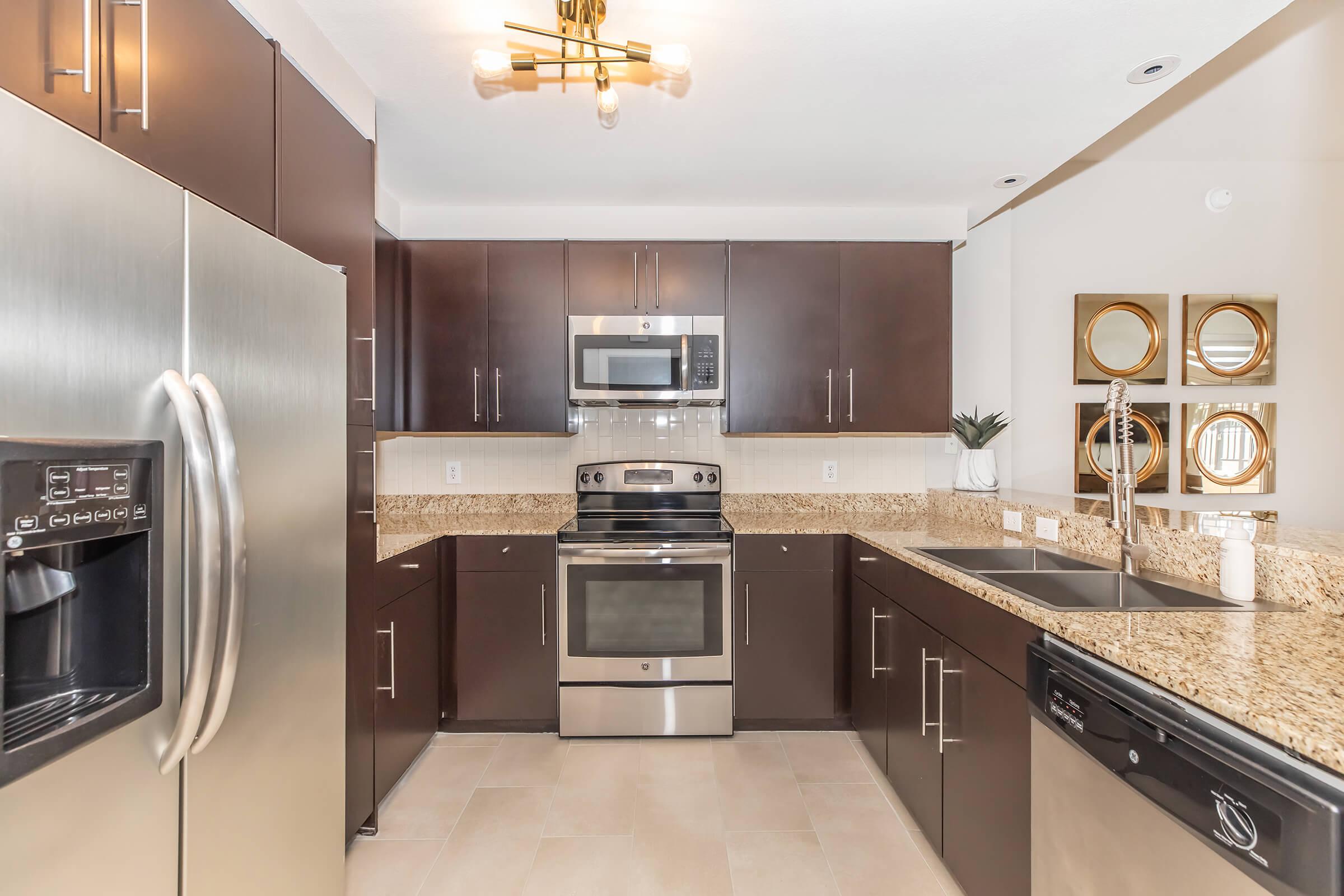 Modern kitchen featuring dark wood cabinetry, stainless steel appliances including a fridge, oven, and microwave, and granite countertops. The space has beige tiled flooring and stylish light fixtures, creating a contemporary and inviting atmosphere. Wall mirrors add a touch of elegance to the design.