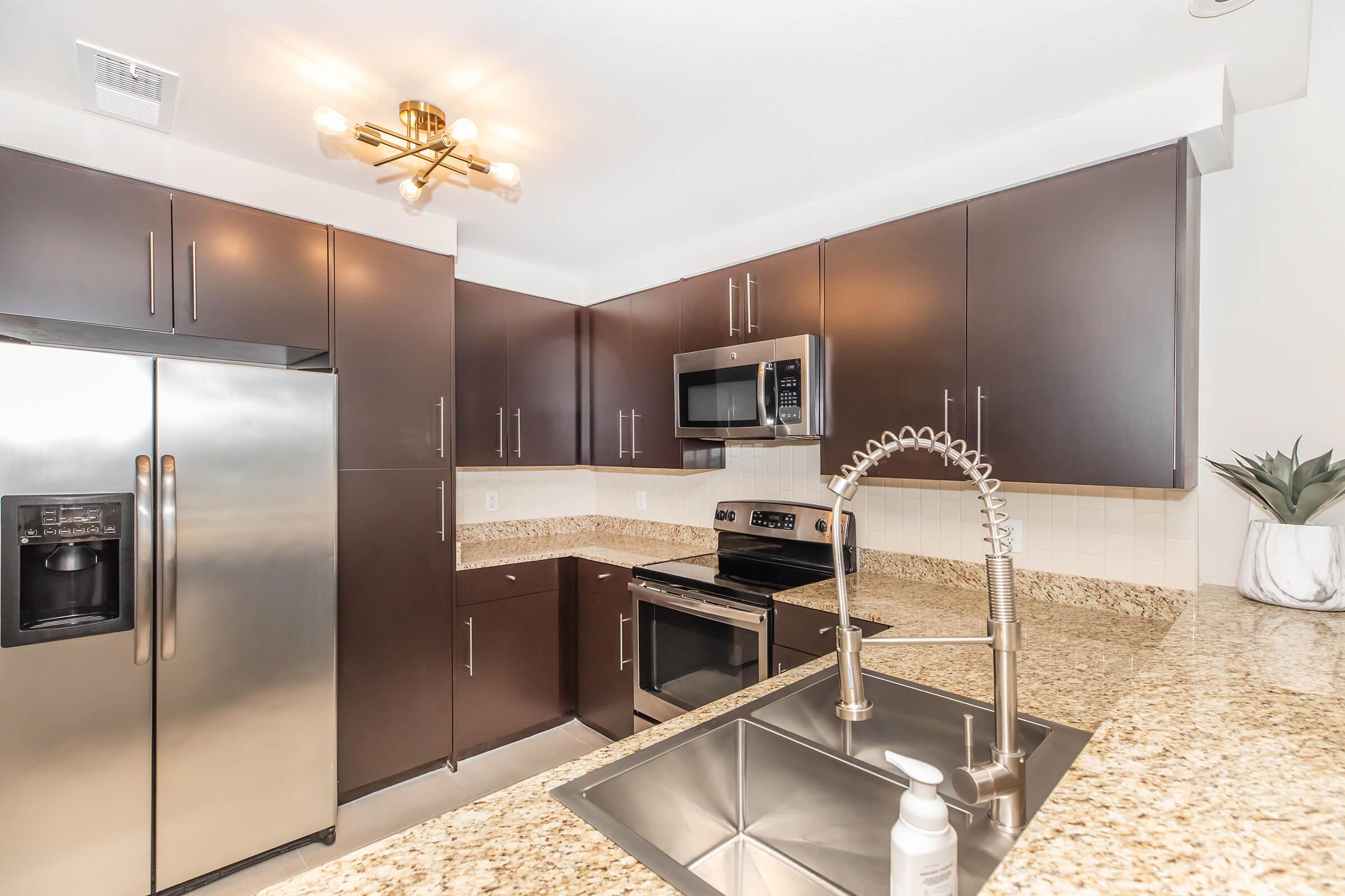 Modern kitchen featuring dark wooden cabinets, stainless steel appliances including a refrigerator and microwave, a granite countertop with a double sink, and a contemporary light fixture. The space is clean and well-lit, emphasizing a stylish and functional design.