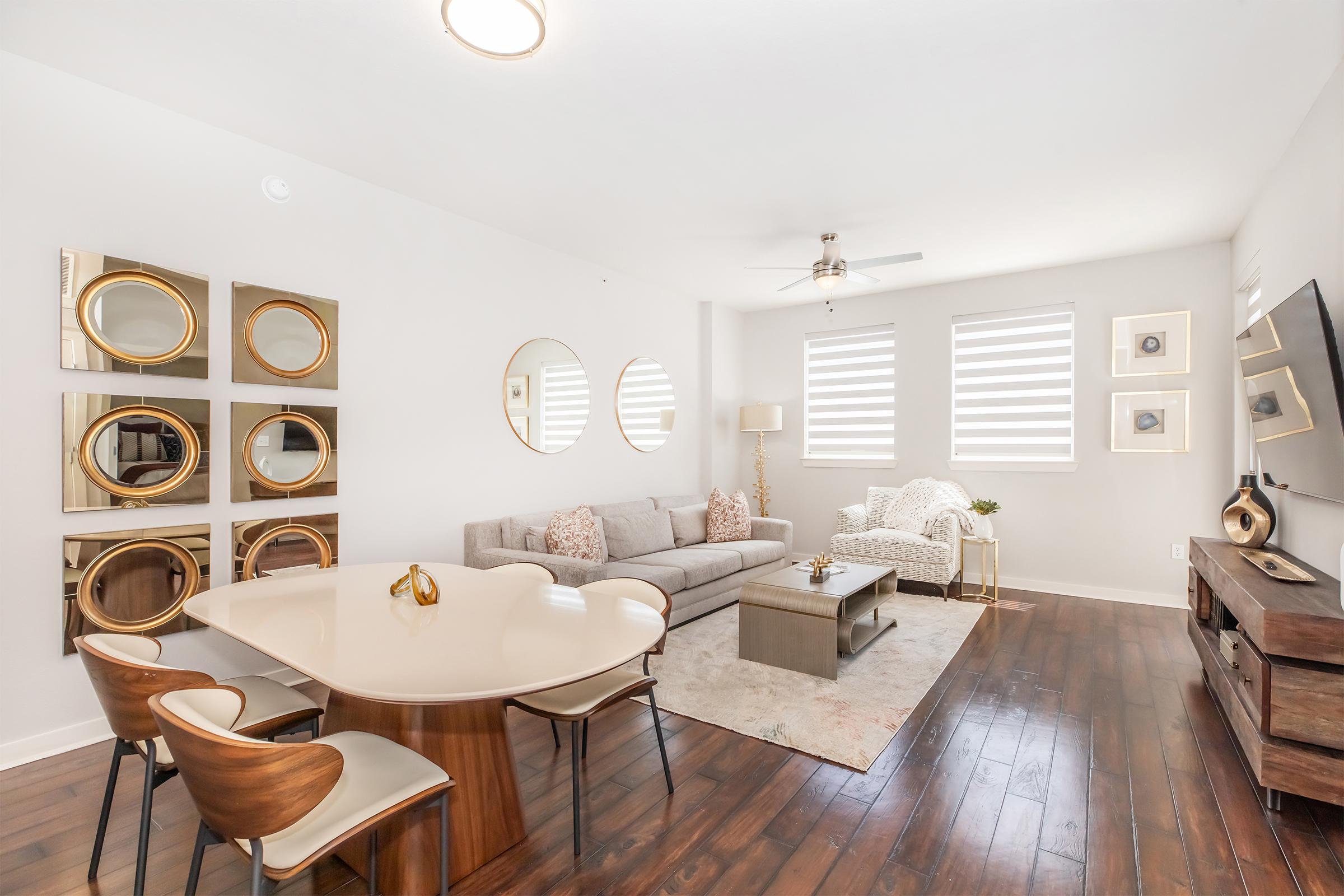 A modern living room featuring a light gray sofa and a cozy armchair. A round dining table with stylish chairs sits nearby. The walls are decorated with circular mirrors, and two windows are covered with striped shades, allowing natural light to fill the space. Hardwood flooring enhances the contemporary look.