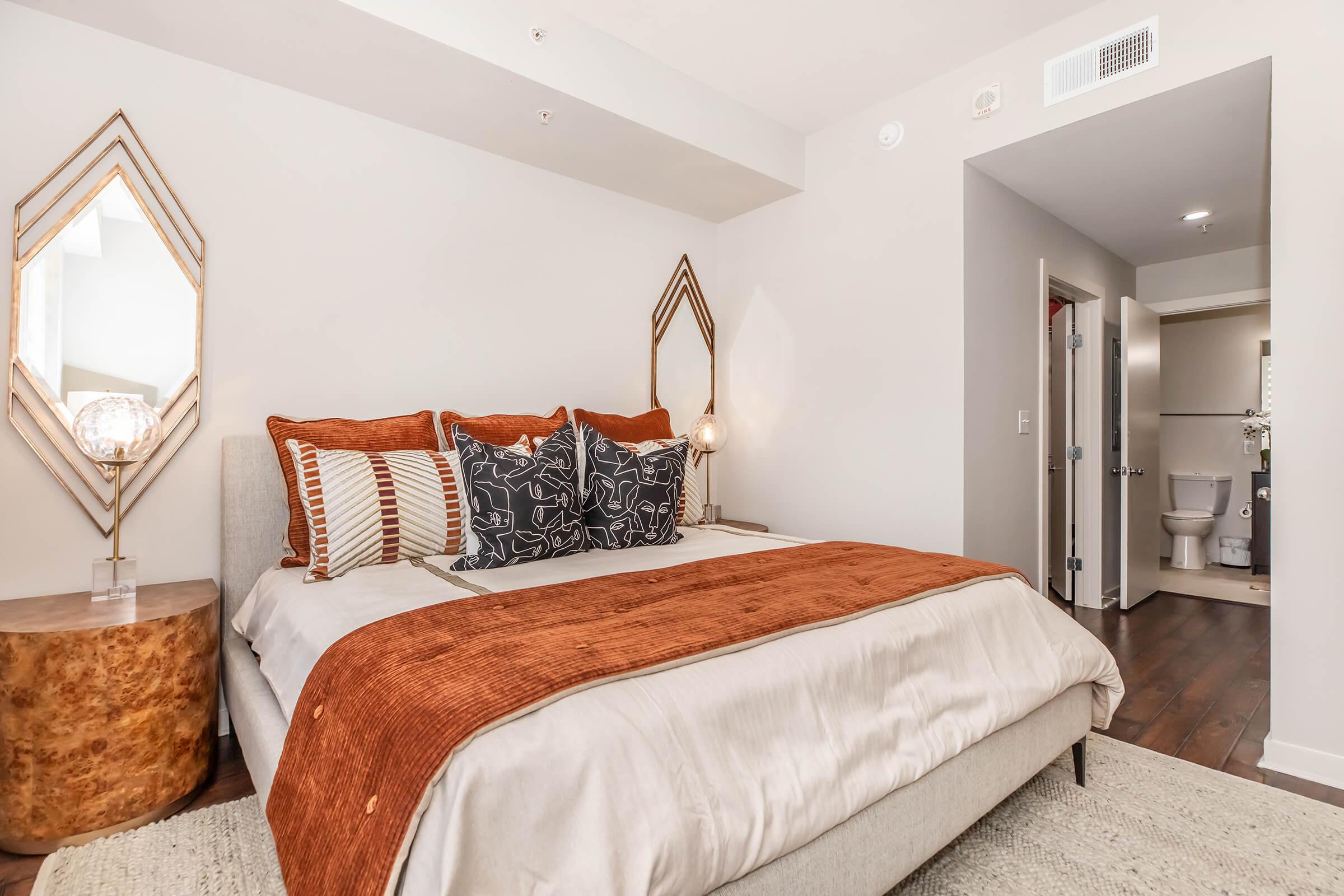 A cozy bedroom featuring a bed with decorative pillows and a warm-colored throw blanket. There is a stylish side table with a lamp, a large mirror on the wall, and a doorway leading to a bathroom. The room has modern decor and wooden flooring, creating a welcoming atmosphere.