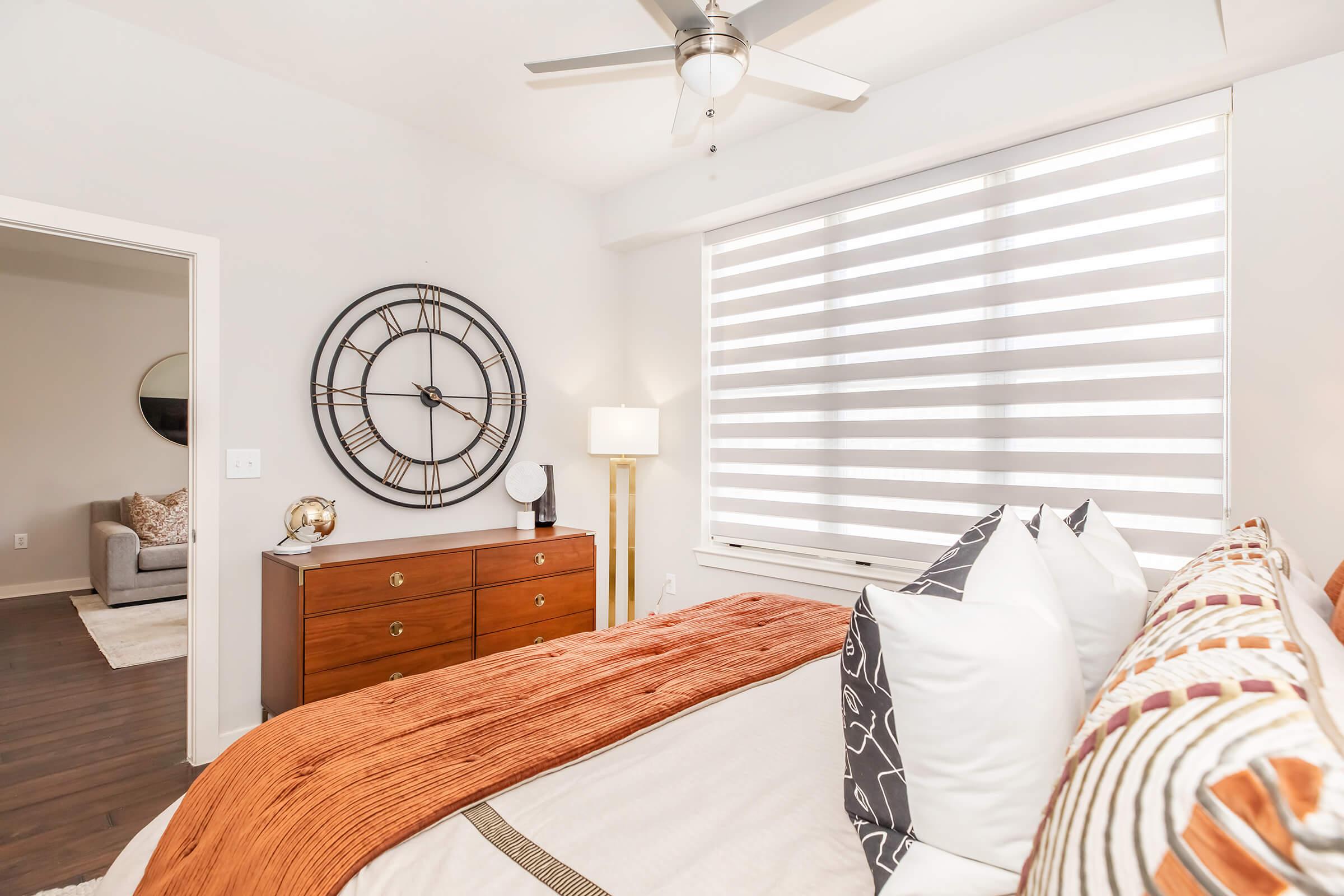 A modern bedroom featuring a neatly made bed with decorative pillows and a rust-colored throw blanket. To the left, a stylish dresser with a large round wall clock above it. A lamp stands beside the dresser, and sheer horizontal blinds cover the window, allowing soft light to enter. The decor is minimalistic and inviting.