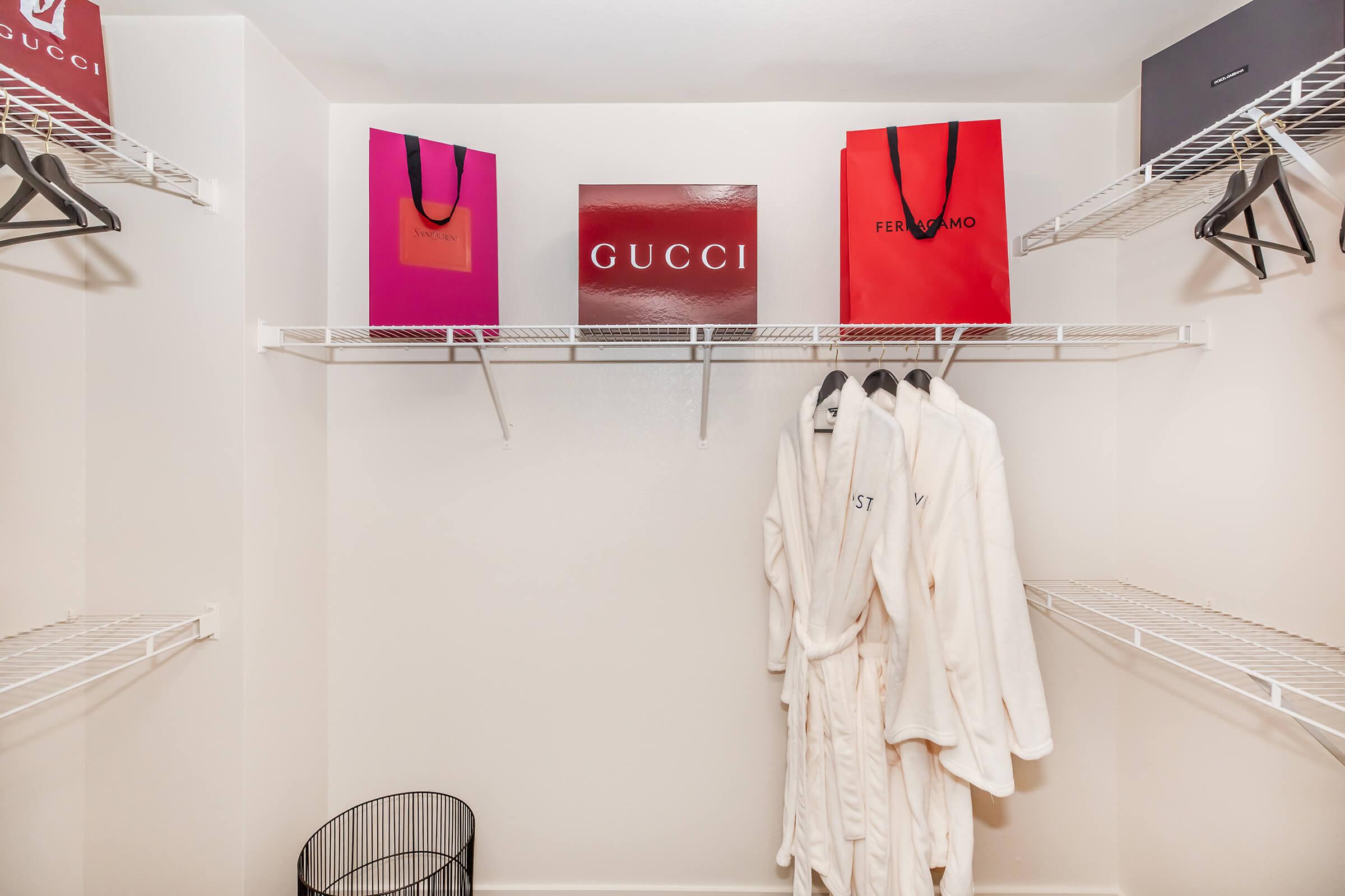 A neatly organized closet featuring luxury brand shopping bags from Gucci and Ferragamo, a large Gucci sign, and white plush bathrobes hanging on wire shelves. A black wire basket is placed at the bottom, adding to the minimalist aesthetic.