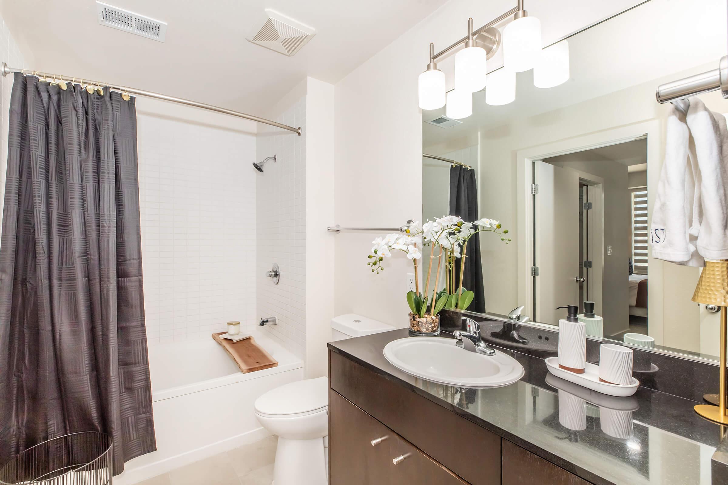 A modern bathroom featuring a bathtub with a shower curtain, a sleek dark wood vanity with a black countertop, and decorative items including a vase with white orchids and toiletries. The space is well-lit with a large mirror and contemporary light fixtures, showcasing a clean and elegant design.