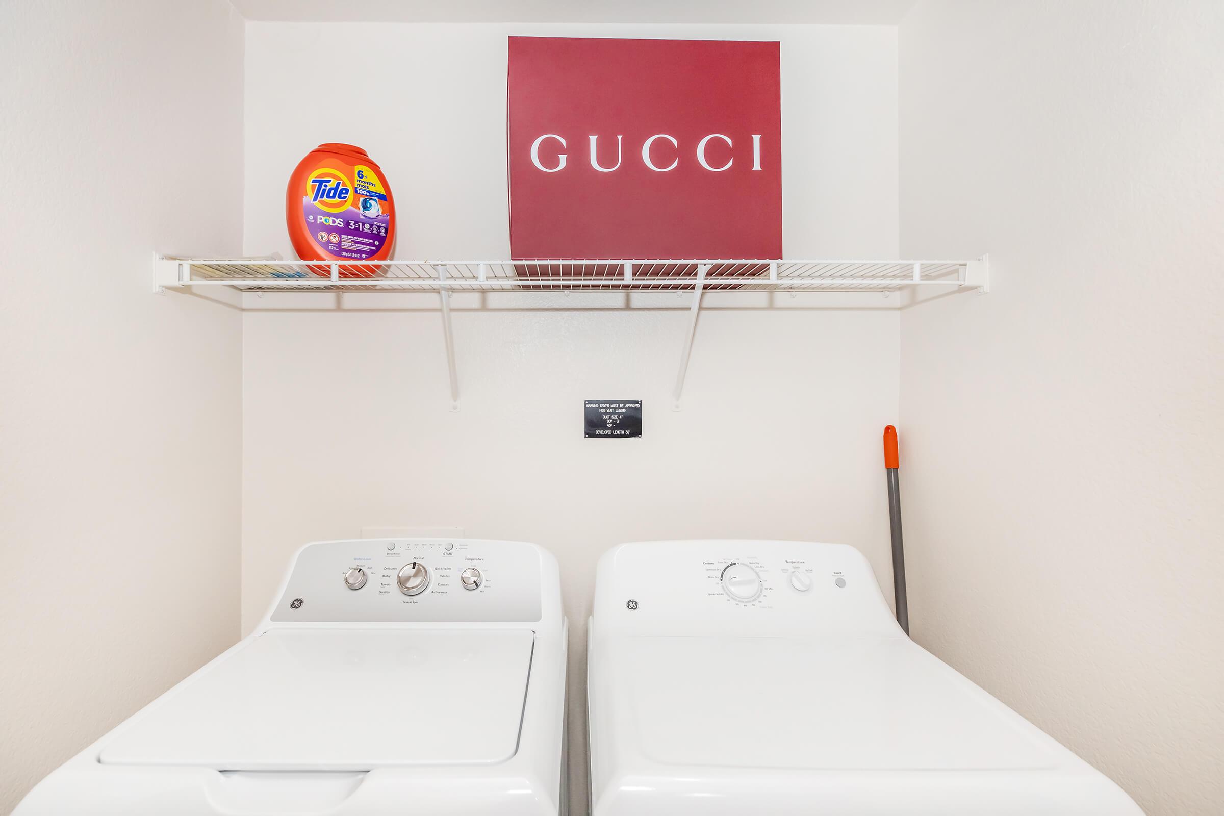 A laundry room featuring a pair of white washing machines neatly positioned side by side. Above them is a shelf holding a bottle of Tide laundry detergent and a red Gucci logo sign against a light-colored wall. A broom can be seen leaning nearby.