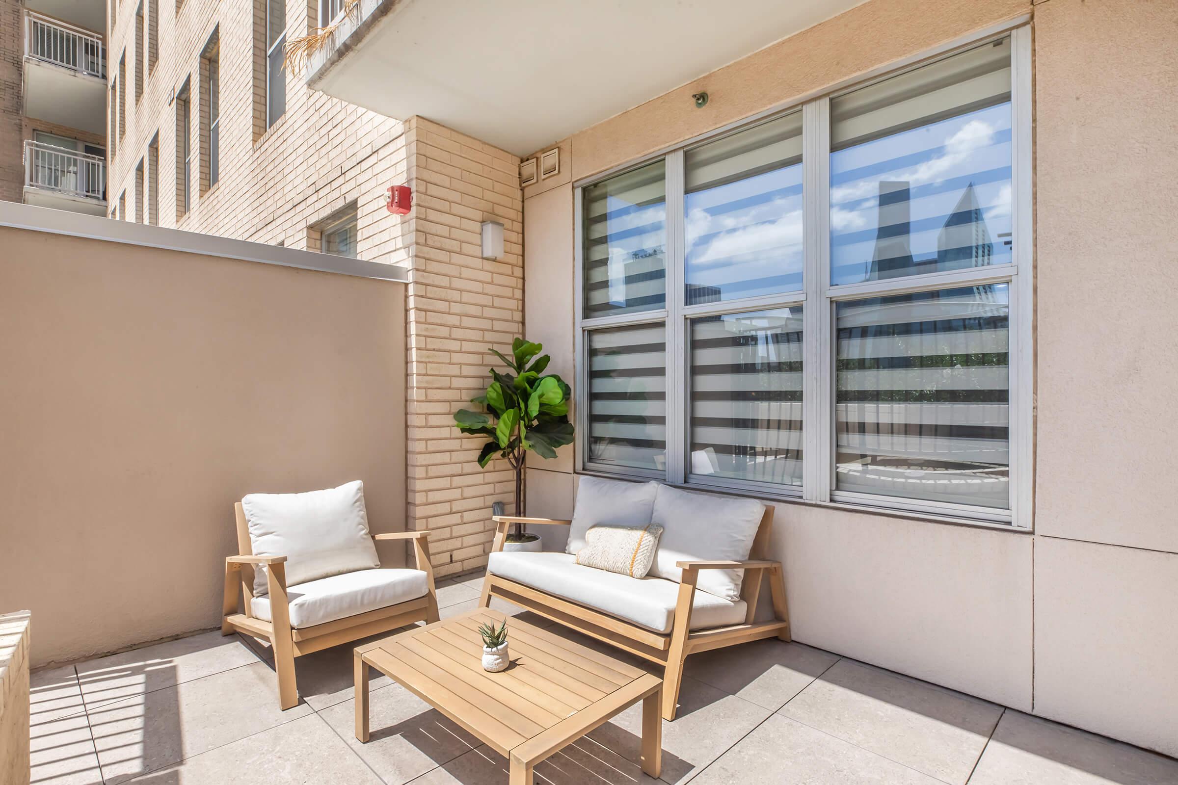 A modern patio with a light-colored sofa and armchair, both featuring white cushions. A small wooden coffee table is in the center, adorned with a potted plant. The patio is enclosed with a wall and has large windows with horizontal blinds, allowing natural light to illuminate the space.