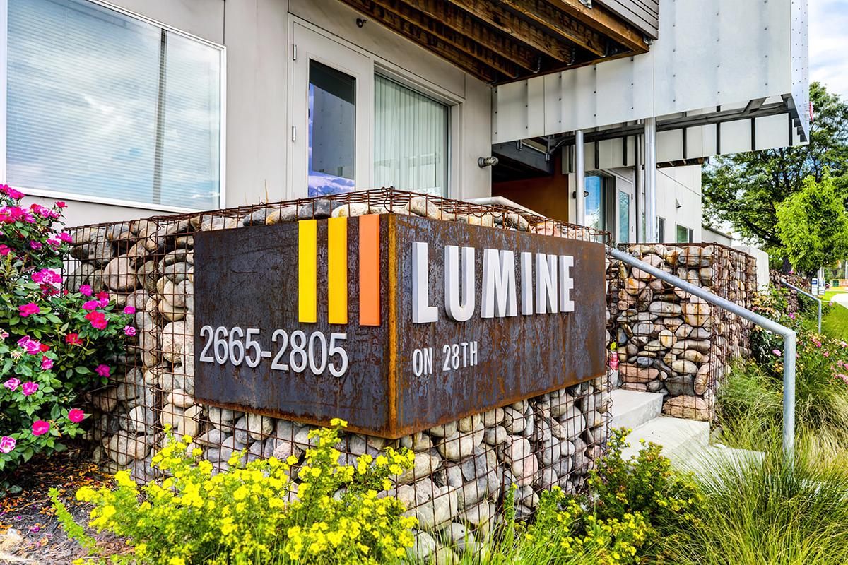 Sign for "Lumine on 28th" featuring the address 2665-2805. The sign is made of rusted metal with bright orange and yellow stripes, surrounded by vibrant flowers and greenery.