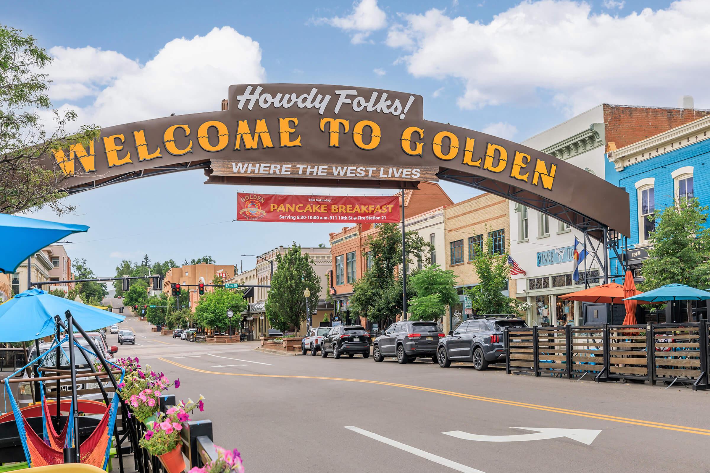 A large welcoming archway that reads "Howdy Folks! Welcome to Golden Where the West Lives" spans across a bustling street lined with trees and colorful outdoor seating. The scene captures a sunny day in a vibrant town, with historic buildings visible in the background.