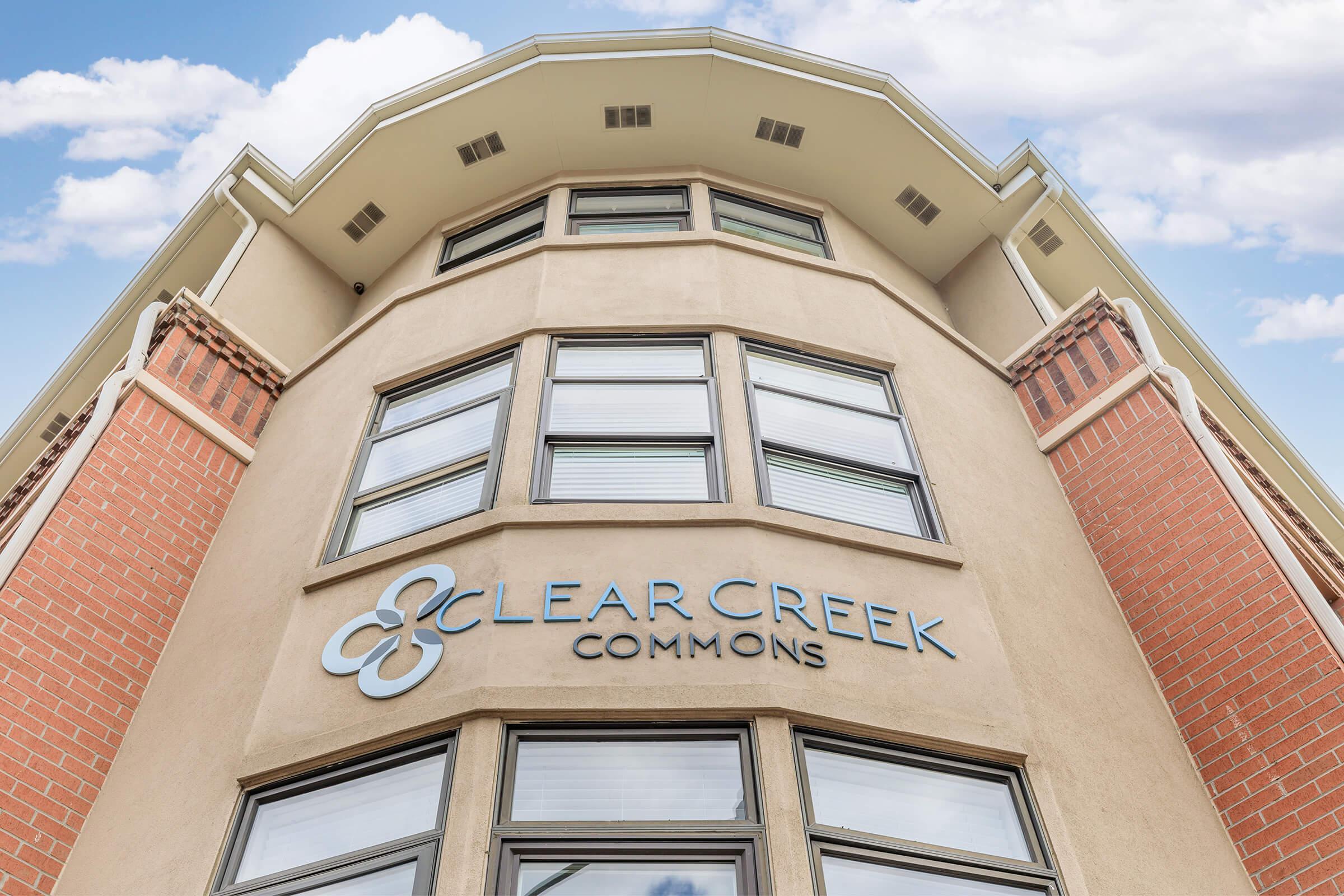A building with a modern architectural design featuring a prominent sign that reads "Clear Creek Commons." The structure has a mix of brick and smooth facade, with multiple large windows reflecting the sky above. Clouds are visible in the background, adding to the aesthetic appeal.