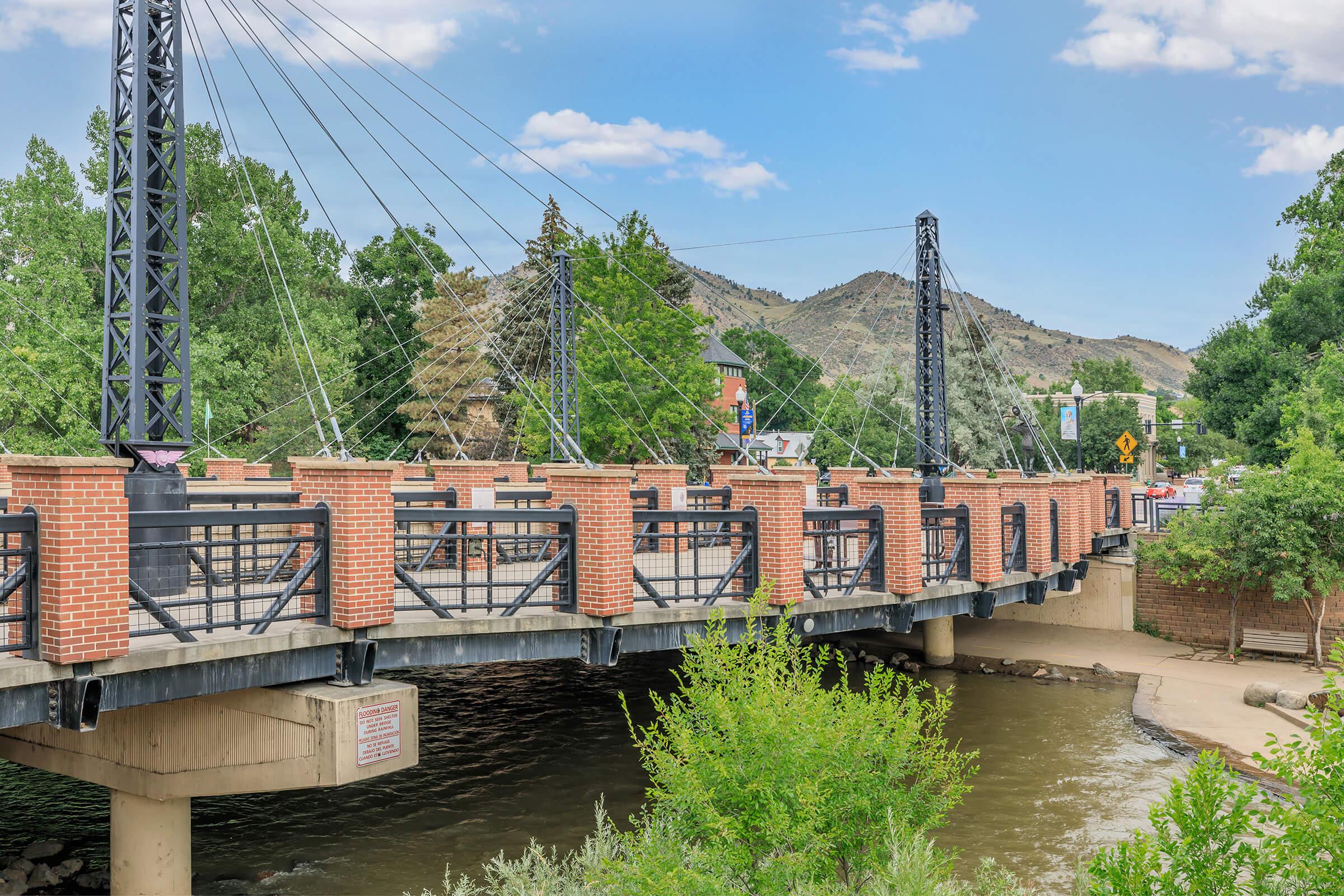 A suspension bridge with brick and metal railings spans a calm river, surrounded by green trees and mountains in the background. The scene is set against a clear blue sky with a few clouds, reflecting a peaceful outdoor environment.
