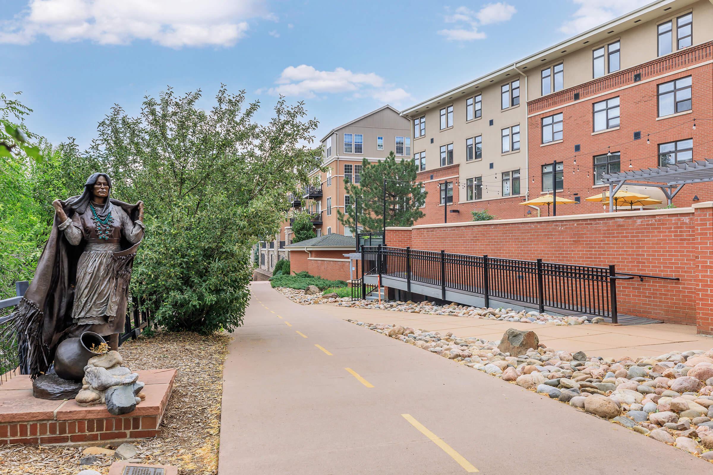 A pathway lined with rocks, leading past a bronze statue of a Native American figure in traditional attire. In the background, modern apartment buildings with balconies are visible, surrounded by greenery and outdoor seating areas. The sky is bright with a few clouds.