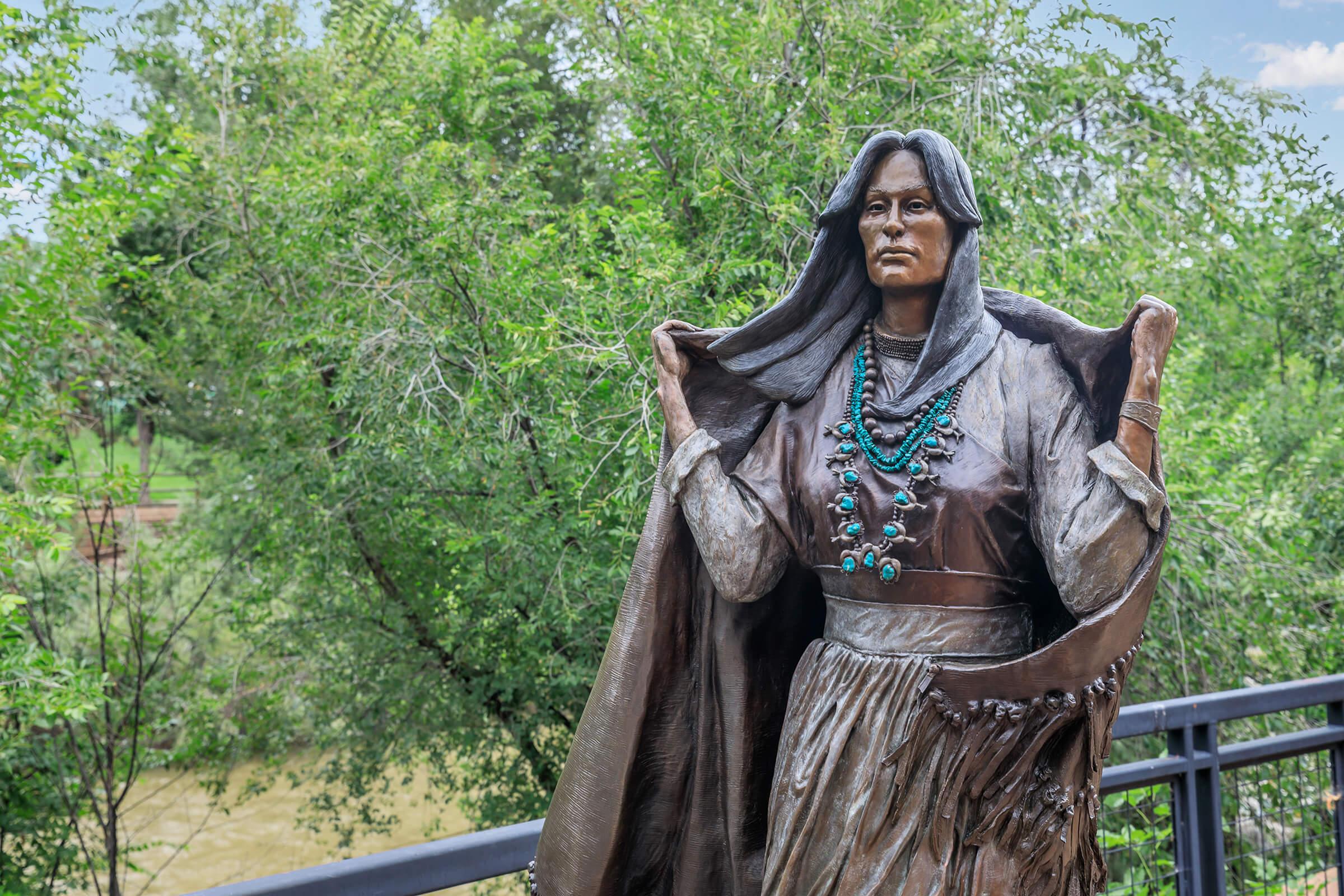 A bronze statue of a Native American woman wearing traditional clothing, adorned with a shawl and turquoise jewelry. The statue is set against a backdrop of green trees and a river, capturing a serene outdoor scene.