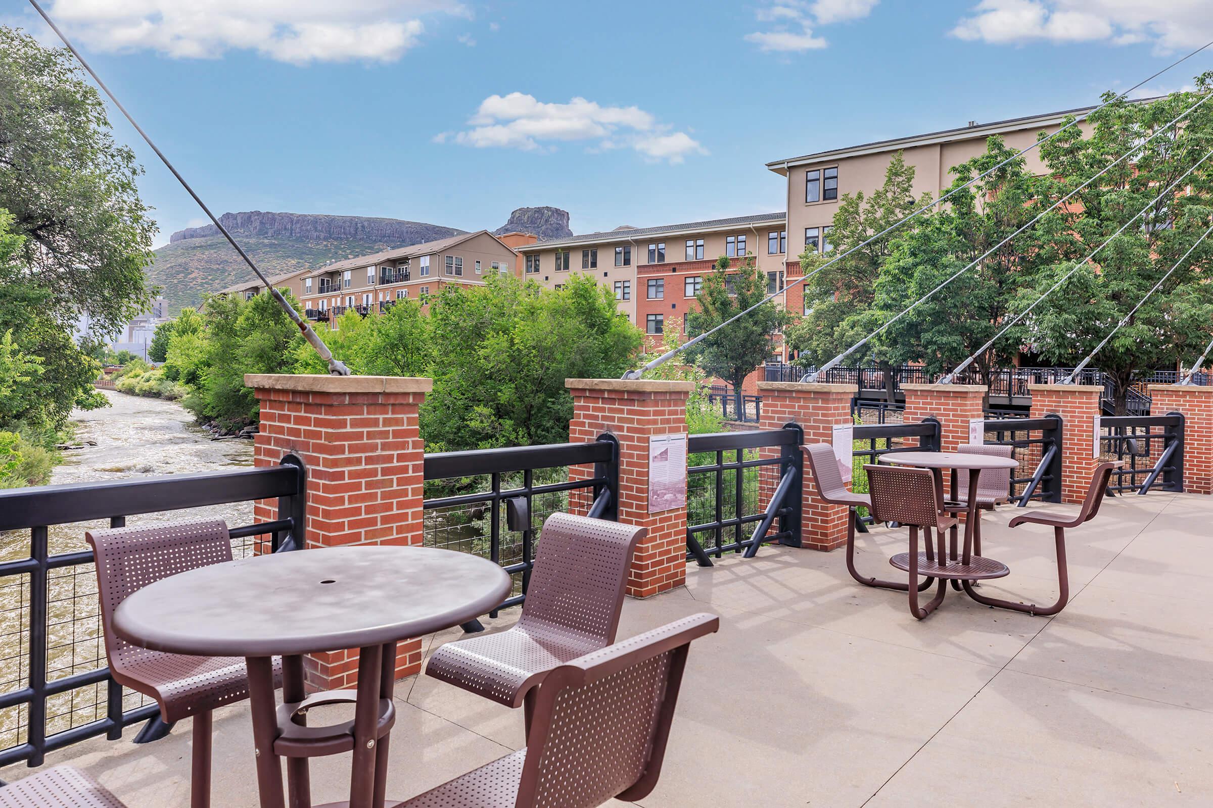 A scenic riverside view featuring a paved walkway with tables and chairs, lush greenery along the water, and modern buildings in the background. The sky is bright with scattered clouds, and a rocky cliff or hill is visible in the distance.