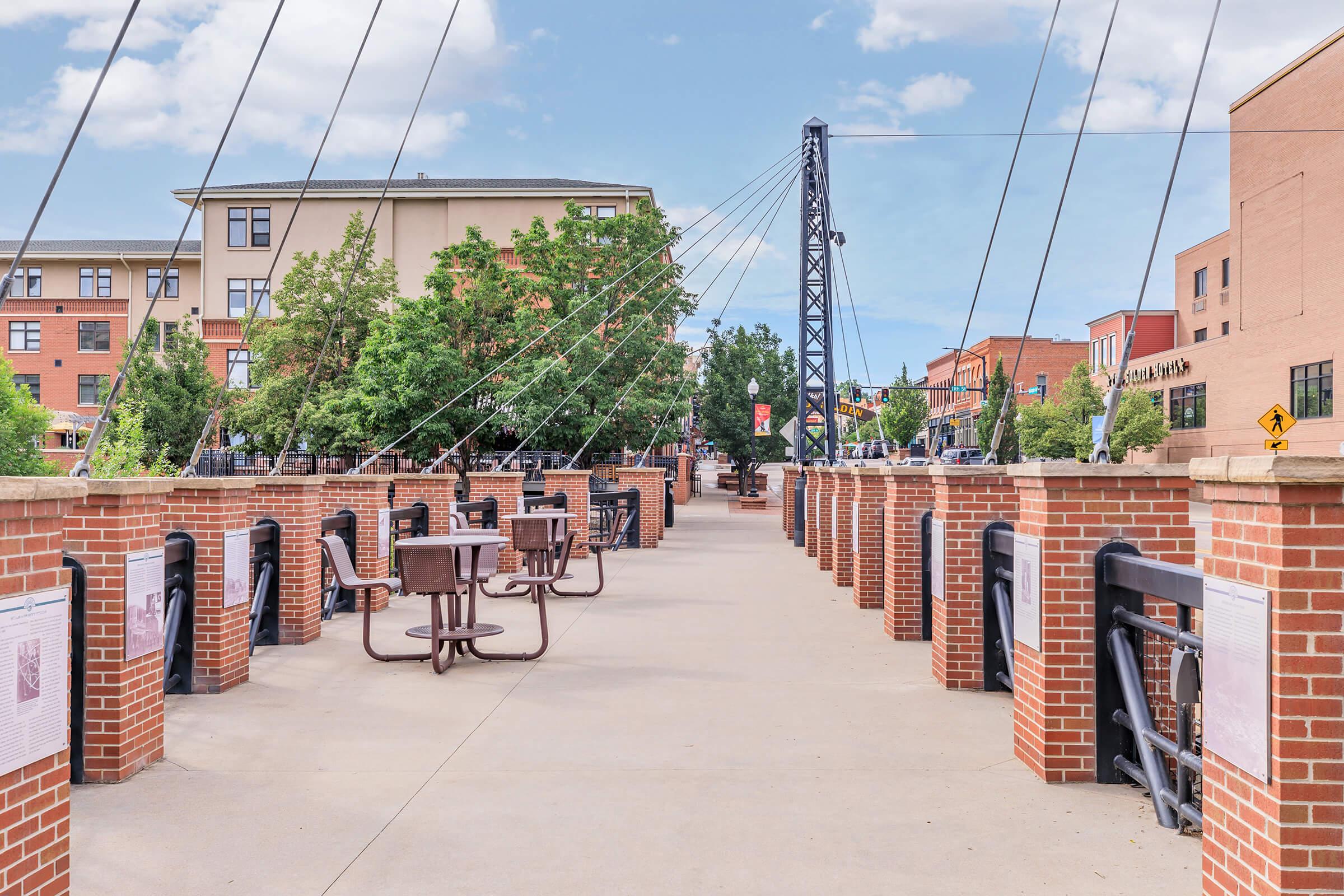 A wide pedestrian walkway showcasing brick-lined paths and tables, flanked by trees and buildings. A large structure with cables towers above, and colorful signs can be seen in the background. The scene is set under a clear blue sky, creating a lively, inviting atmosphere.
