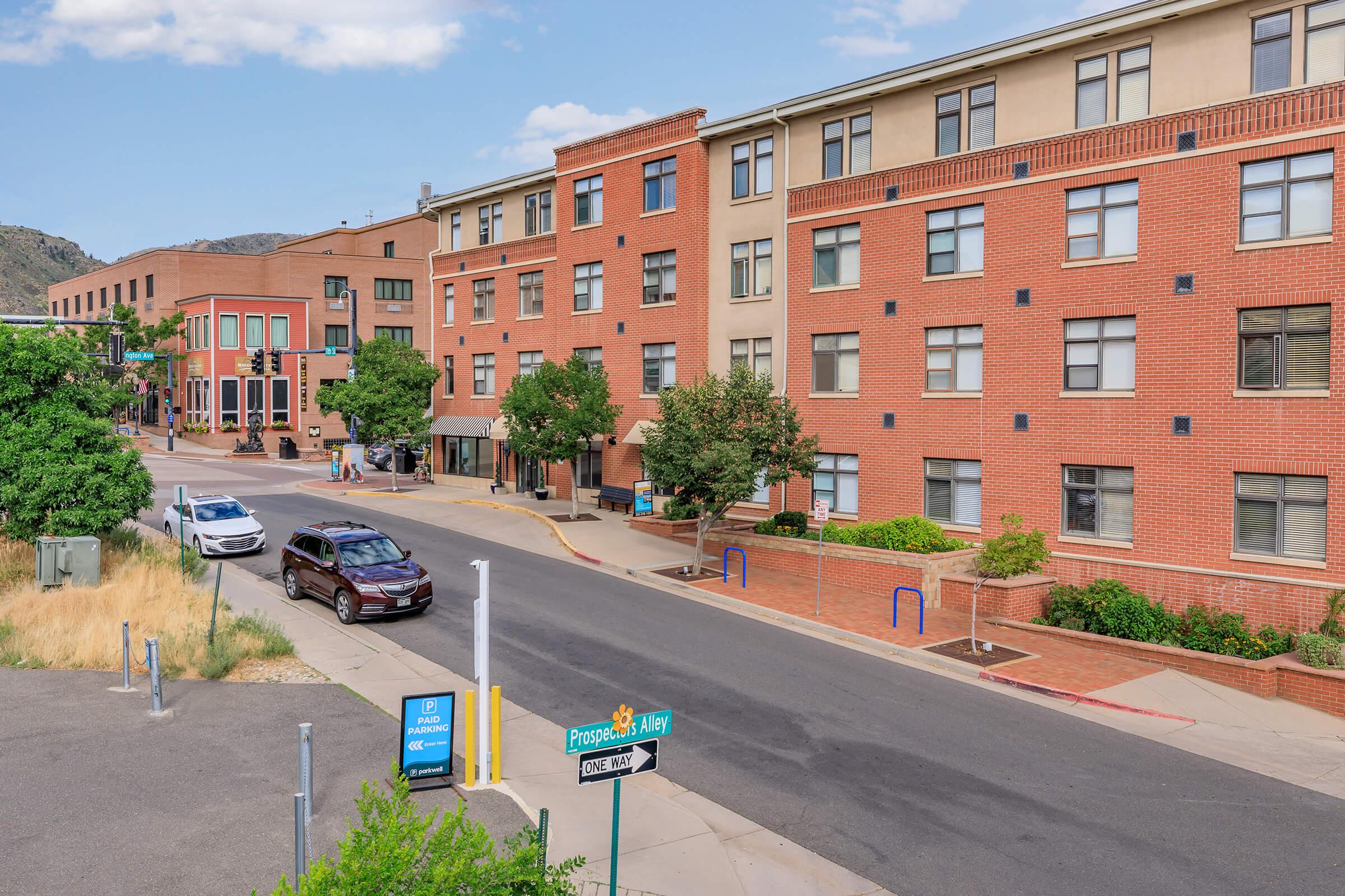 A view of a residential area with modern brick buildings lining a street. There are trees and parking spaces visible, along with a street sign for "Prospect Alley." The scene is set in a sunny environment with clear skies and mountains in the background.