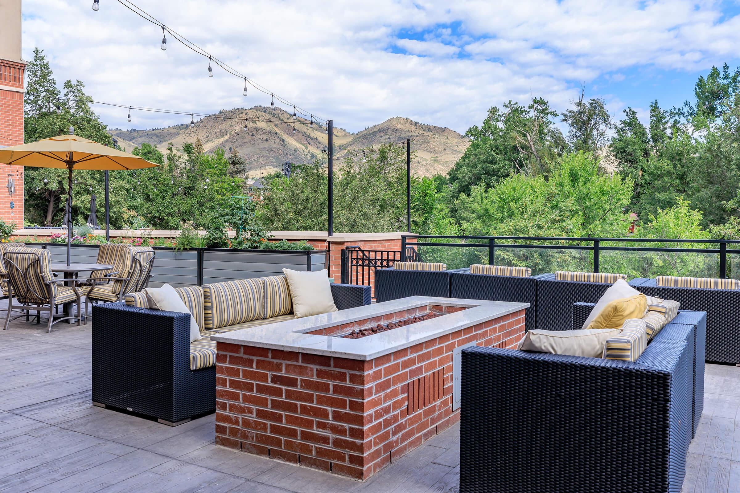 A modern outdoor lounge area featuring comfortable seating with striped cushions, a fire pit made of brick, and an umbrella. The backdrop includes rolling hills and trees under a partly cloudy sky, creating a tranquil atmosphere for relaxation.