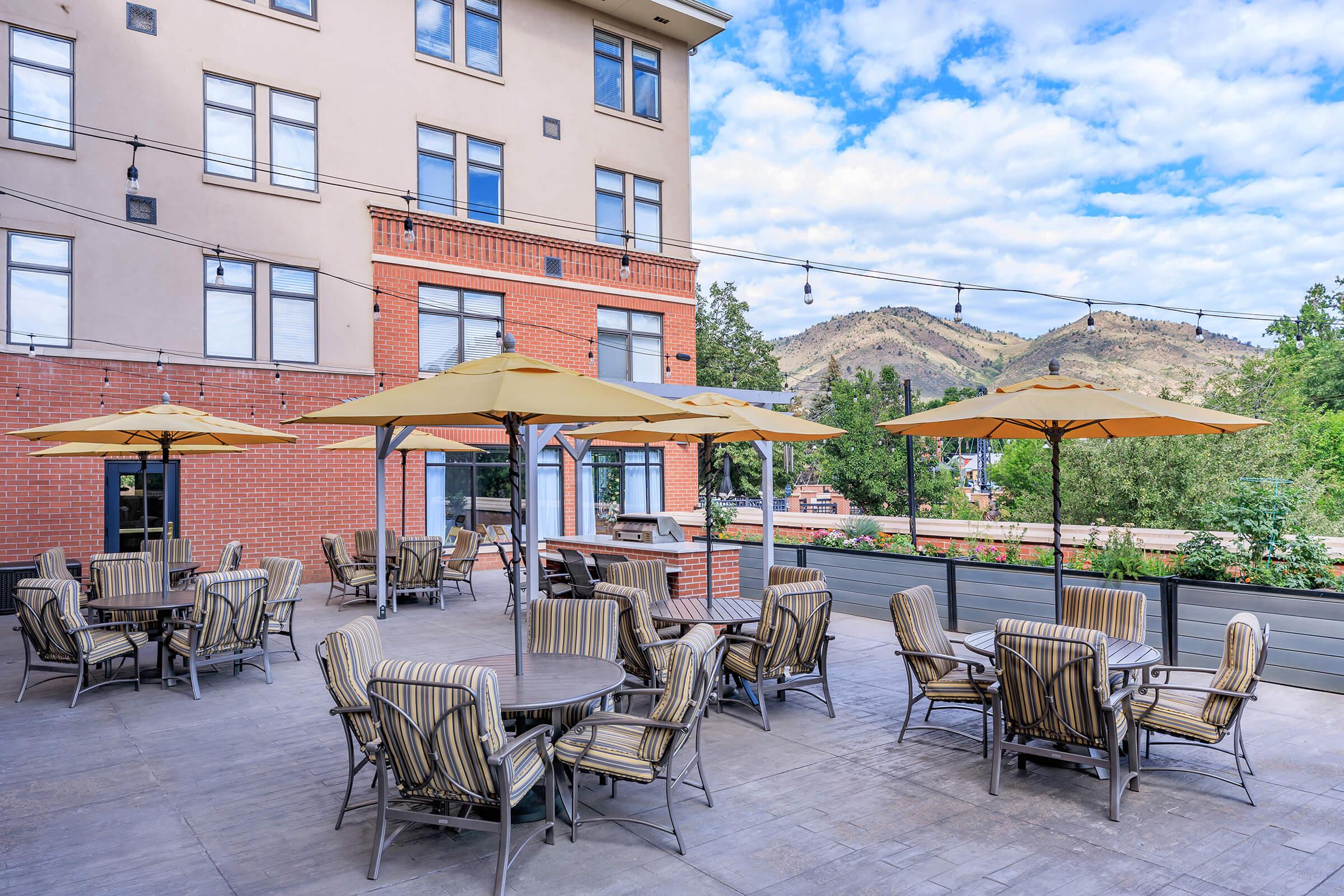 Outdoor seating area featuring several patio tables and chairs with striped umbrellas. The backdrop includes a building with large windows and a view of green hills and blue sky with scattered clouds. The setting is ideal for social gatherings or dining al fresco.