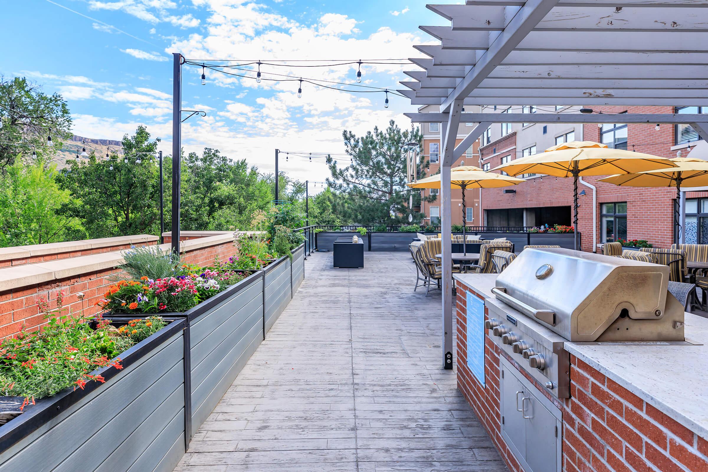 A modern outdoor patio with a wooden deck, featuring a stainless steel grill, planters with colorful flowers, and tables with yellow umbrellas. String lights are hung overhead, and greenery surrounds the area, providing a pleasant, inviting atmosphere for gatherings and relaxation.