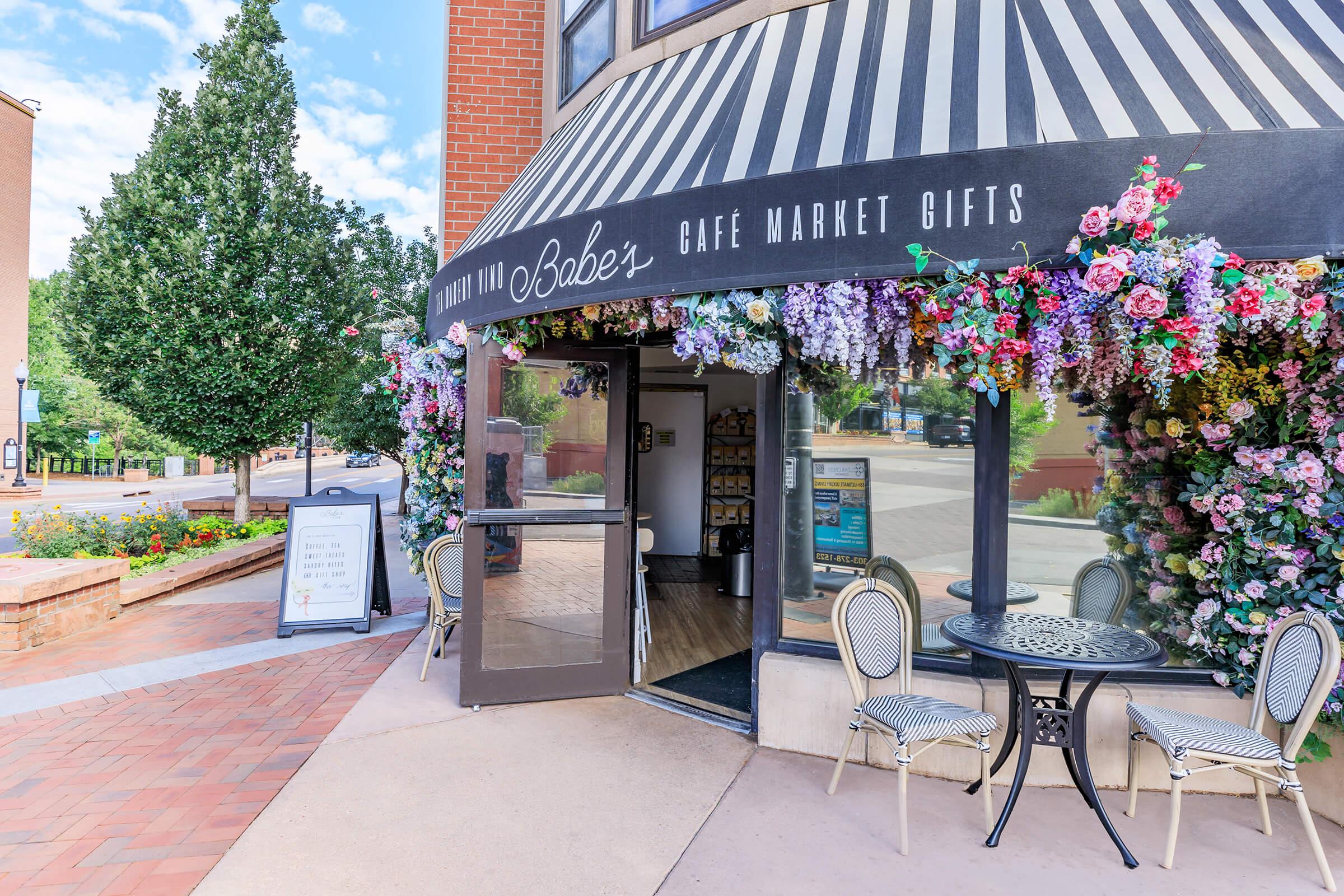 Entrance to Babe's Cafe Market Gifts, featuring a striped awning adorned with colorful flowers. Two outdoor chairs are set at a small table, with a sign nearby. The surrounding area includes trees and a brick walkway, creating a welcoming atmosphere.
