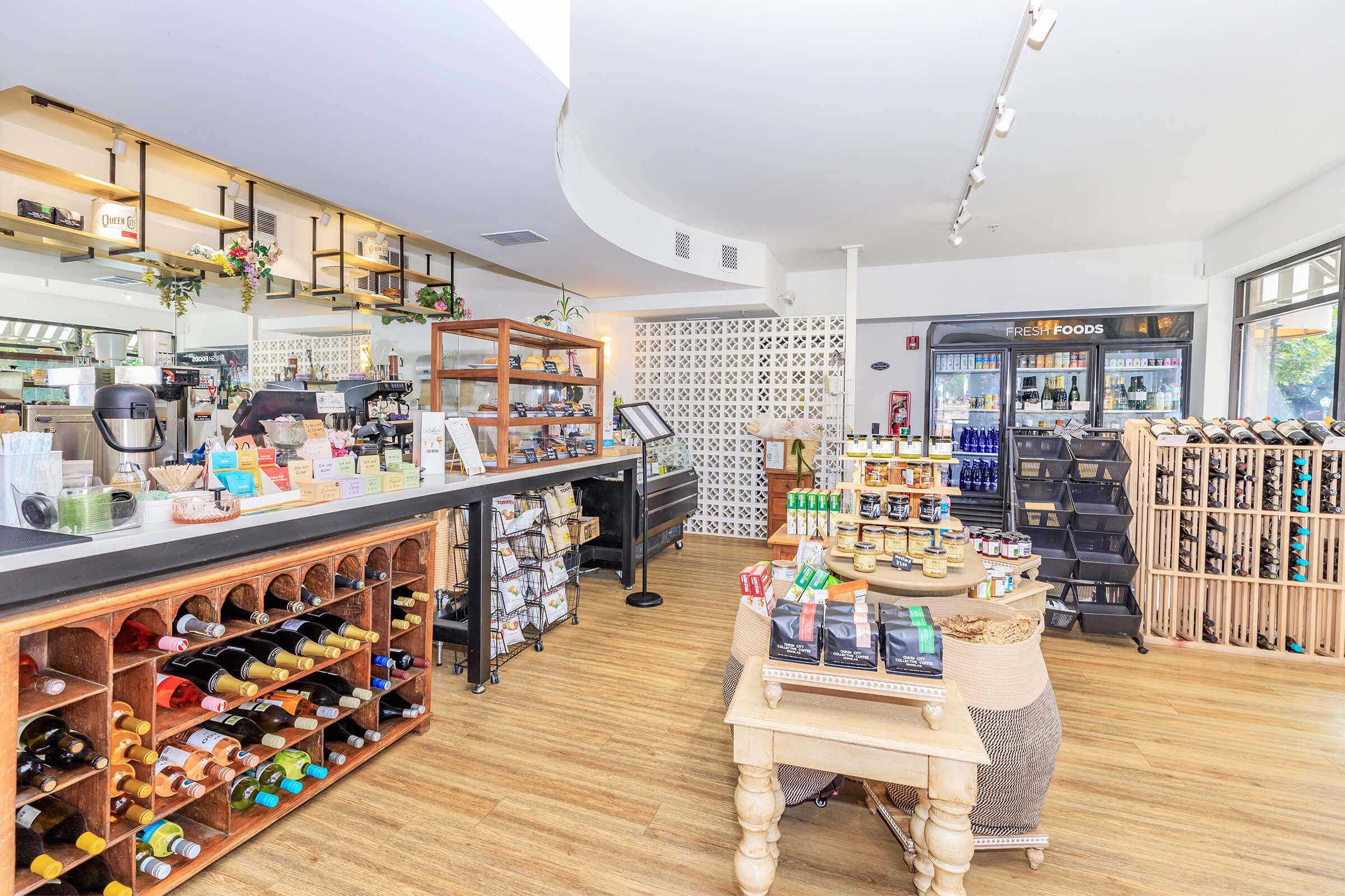 Interior of a modern store featuring a coffee bar, shelves with various beverages and snacks, and a display of wines. The floor is wooden, and there are product displays arranged neatly, with natural lighting illuminating the space.