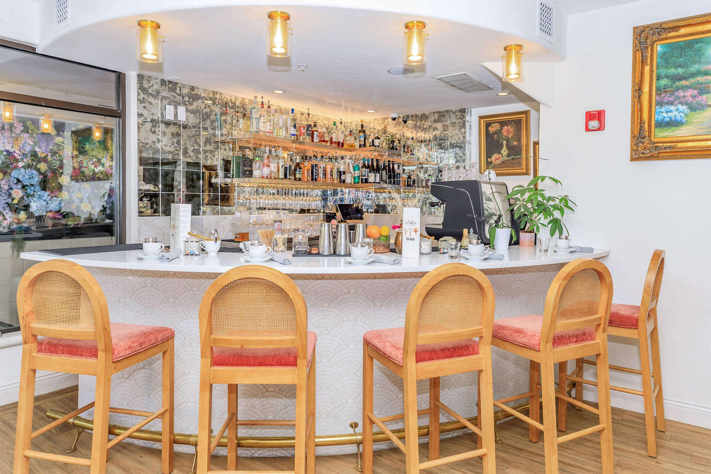 A modern bar area featuring a curved counter with high wooden stools, adorned with pink cushions. Behind the bar, shelves showcase a variety of spirits and glassware. The ambiance is bright and inviting, complemented by decorative plants and framed artwork on the walls.