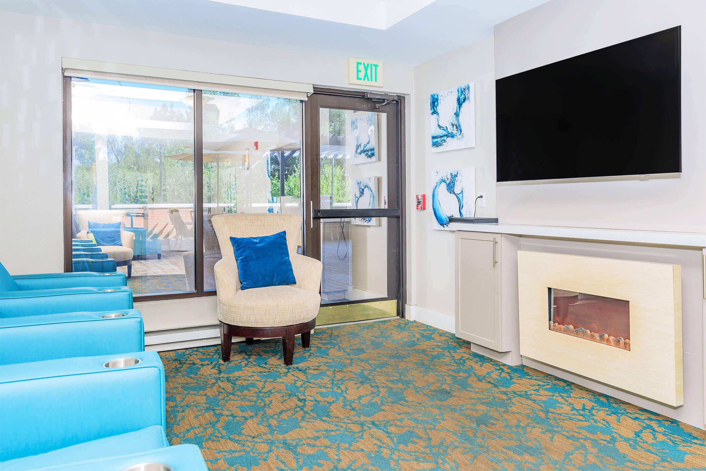 A cozy indoor lounge area featuring a light blue armchair with a blue pillow, a flat-screen TV mounted on the wall, a modern electric fireplace, and a glass door leading to an outdoor space. The carpet has a decorative pattern, and there are blue and white wall art pieces.