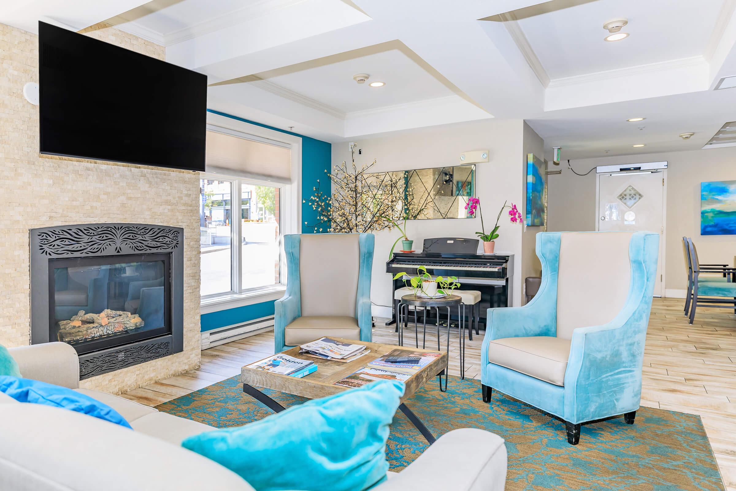 A cozy lounge area featuring two upholstered blue armchairs and a light-colored sofa. A wooden coffee table is in the center, surrounded by magazines. A modern fireplace is mounted on a textured wall, and a piano with decorative flowers adds elegance. Soft lighting enhances the inviting atmosphere.
