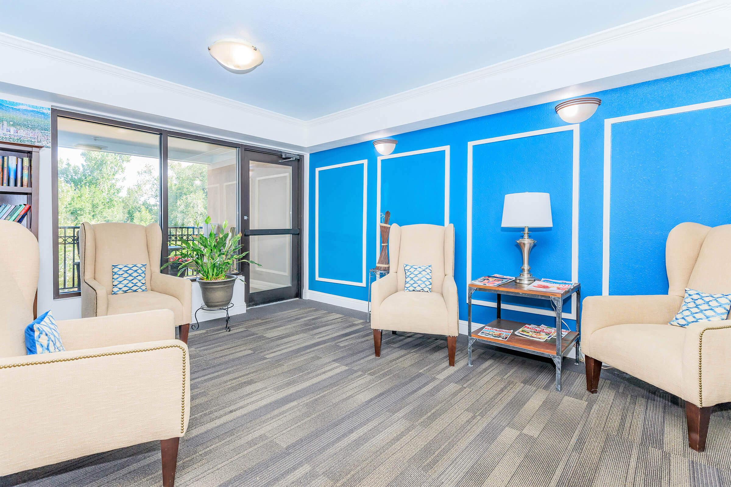A cozy lounge area featuring two beige wingback chairs with blue accent pillows on either side of a small table. A tall indoor plant adds greenery, while a bookshelf and magazines are visible on a side table. The walls are painted bright blue with white trim, enhanced by natural light from large glass doors.