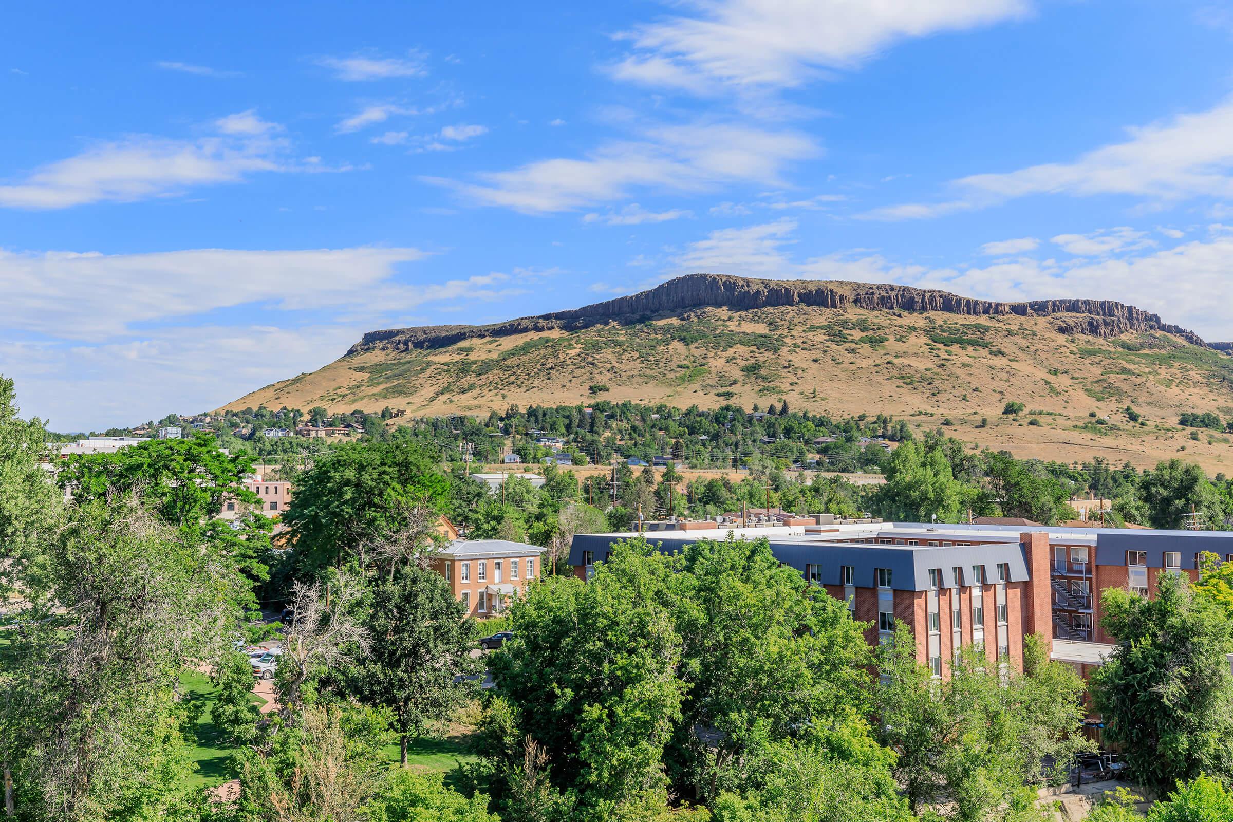 A scenic view of a mountainous landscape featuring a flat-topped hill in the background. In the foreground, there are several buildings surrounded by lush green trees, with a clear blue sky scattered with clouds above. The scene reflects a peaceful and natural environment.