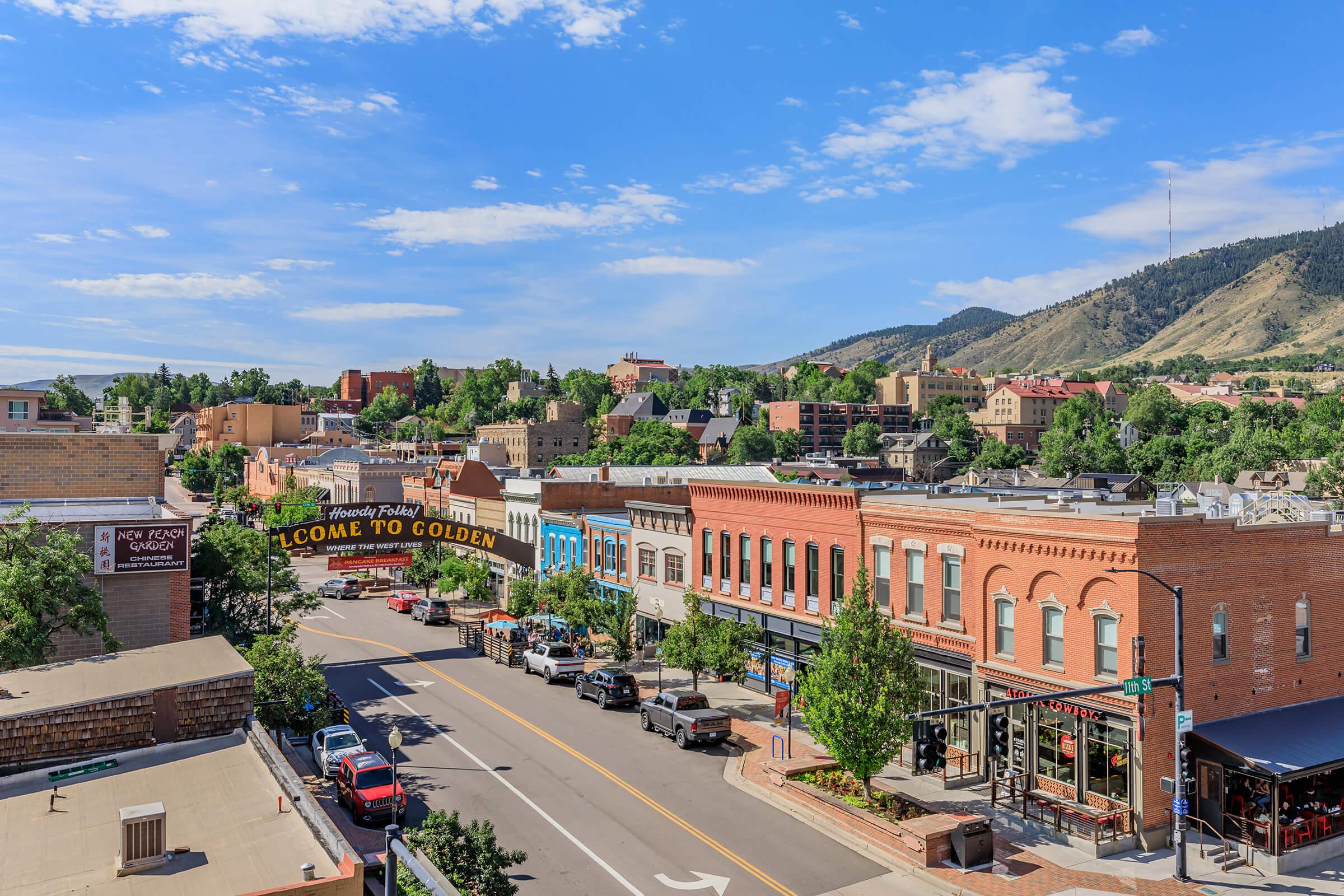 A vibrant view of a small town with historic buildings, colorful storefronts, and trees lining the street. A welcoming sign reads "Welcome to Golden." In the background, rolling hills and blue sky complete the picturesque scene, capturing the charm of the area.